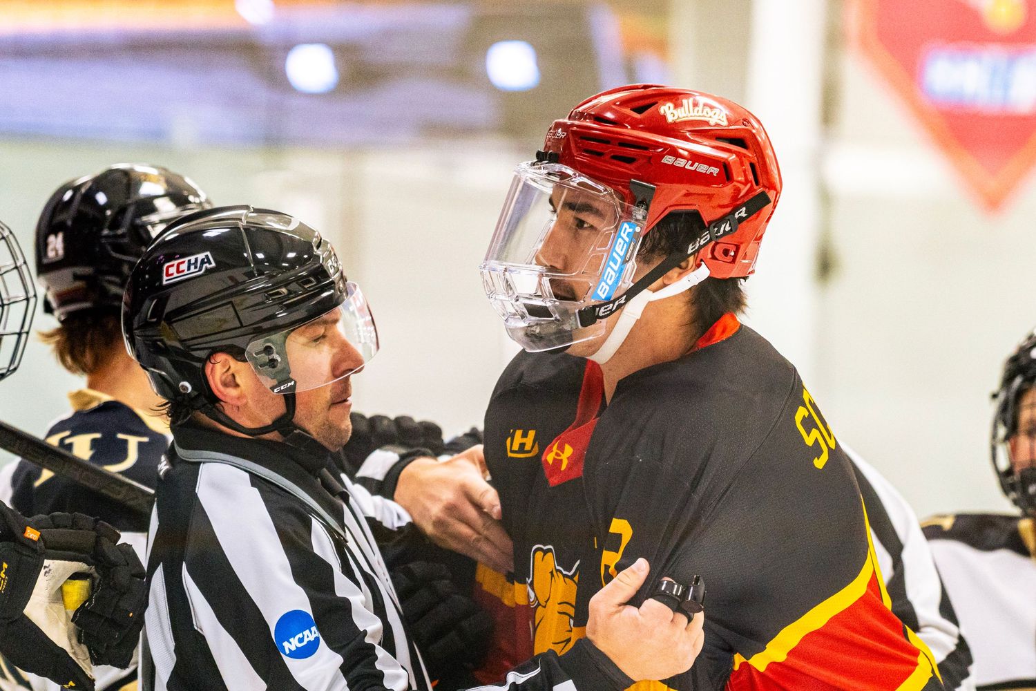 Hockey referee in a striped jersey and black helmet talks with a player in a red helmet and black and gold jersey during an ice hockey game.