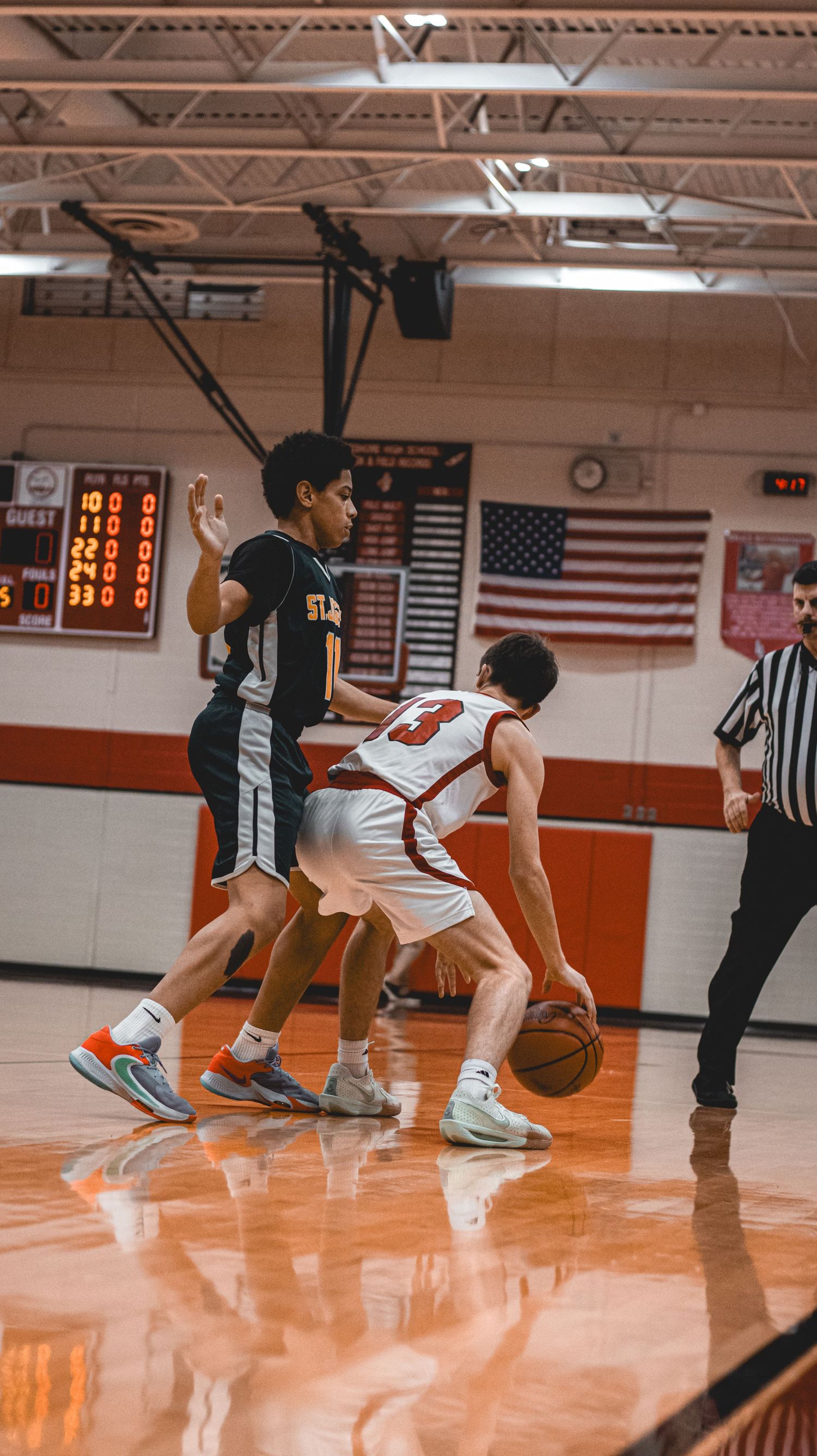 Basketball players compete on shiny gymnasium floor with American flag visible in background.