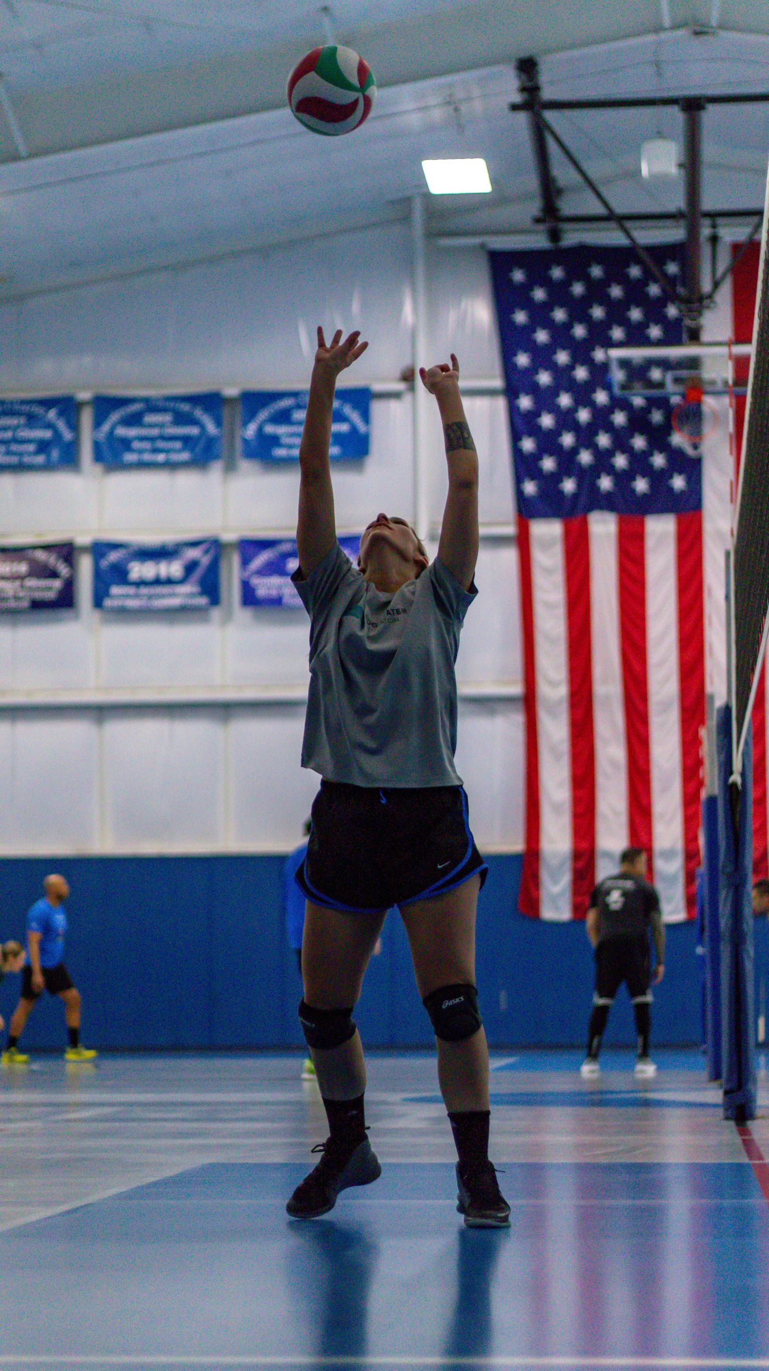 Volleyball player setting the ball in front of an American flag in an indoor gymnasium.