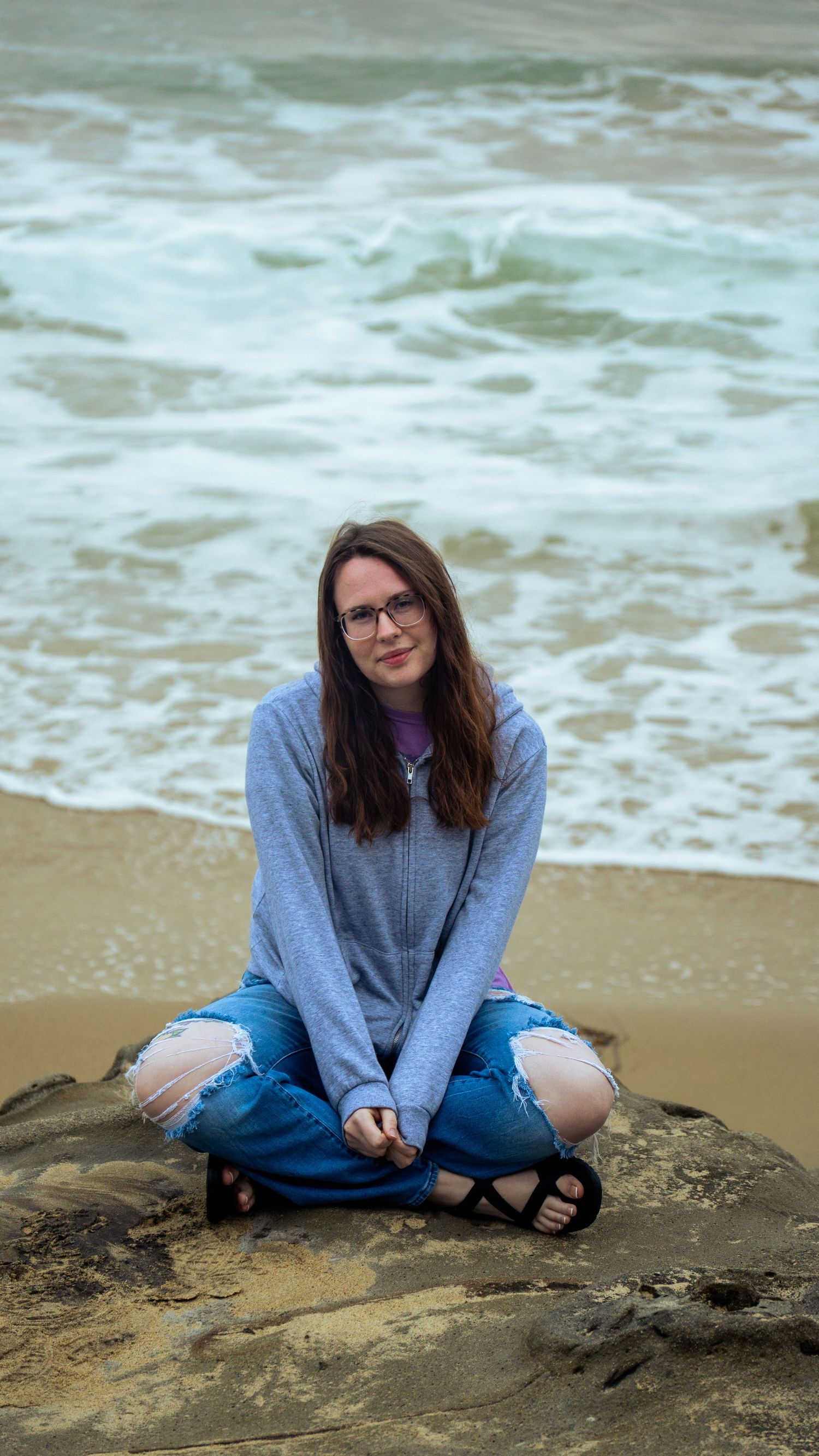 Person in glasses and blue sweater sitting cross-legged on rocky beach with ocean waves in background.