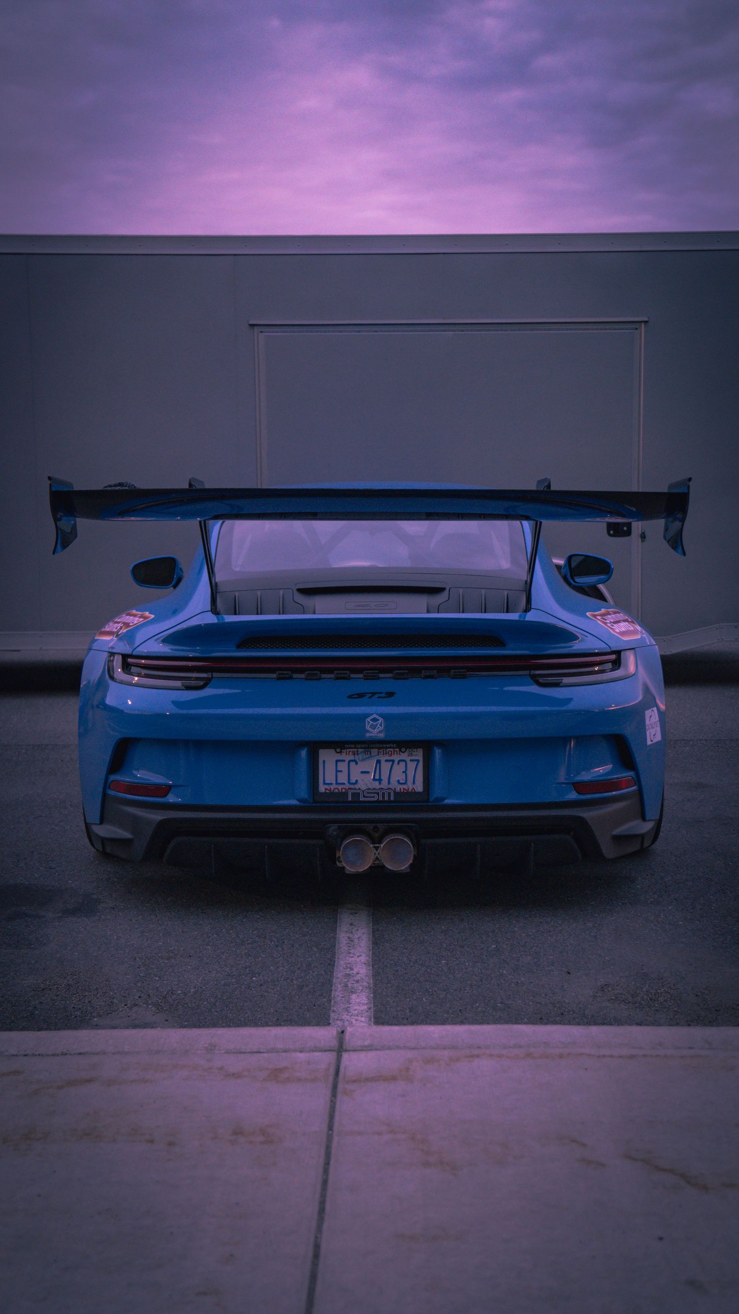 Rear view of blue Porsche GT3 with large rear wing against purple twilight sky.
