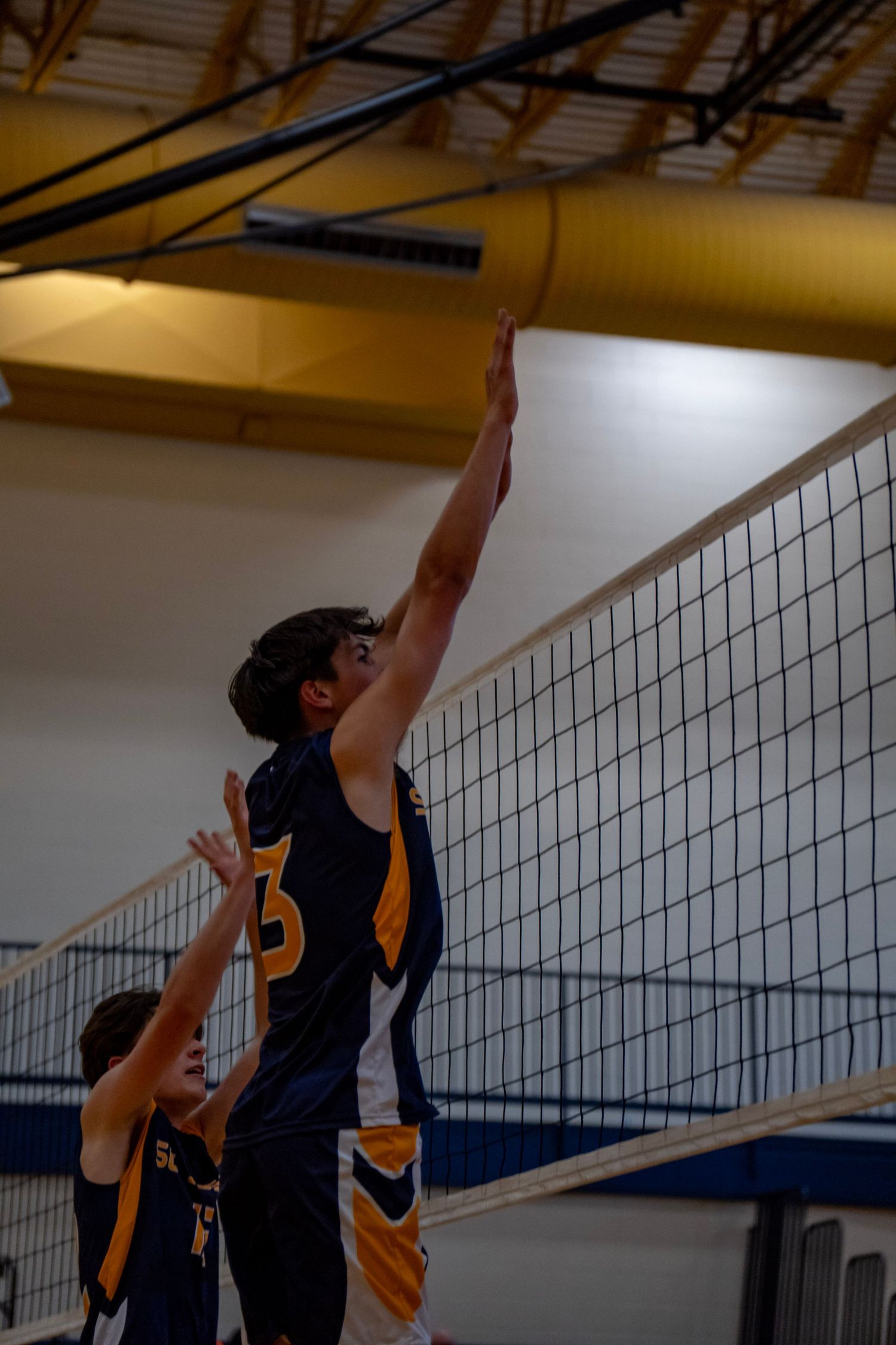 Player in navy uniform reaches up for block at volleyball net during game.
