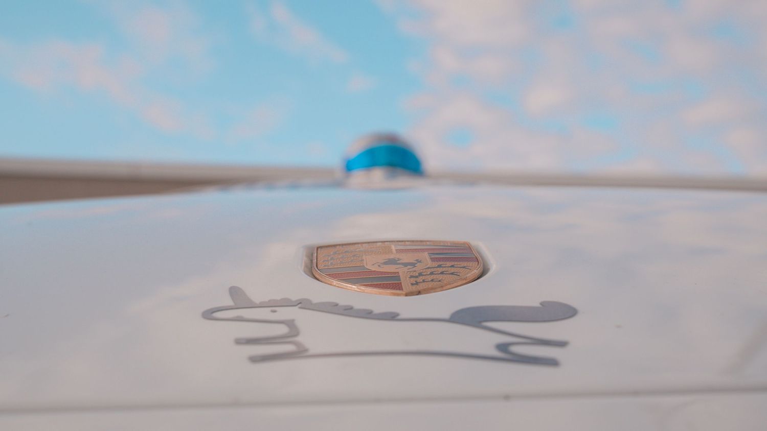 White car hood with Porsche emblem and blurred blue element visible in background.
