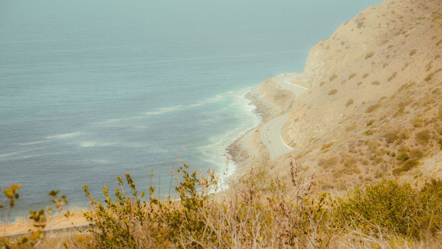 Rolling coastal hills with golden vegetation overlooking a pristine beach and blue ocean waters.