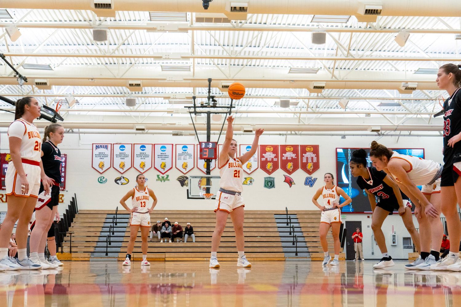 Basketball players compete for a rebound during an intense game in a high school gymnasium with banners on the walls.