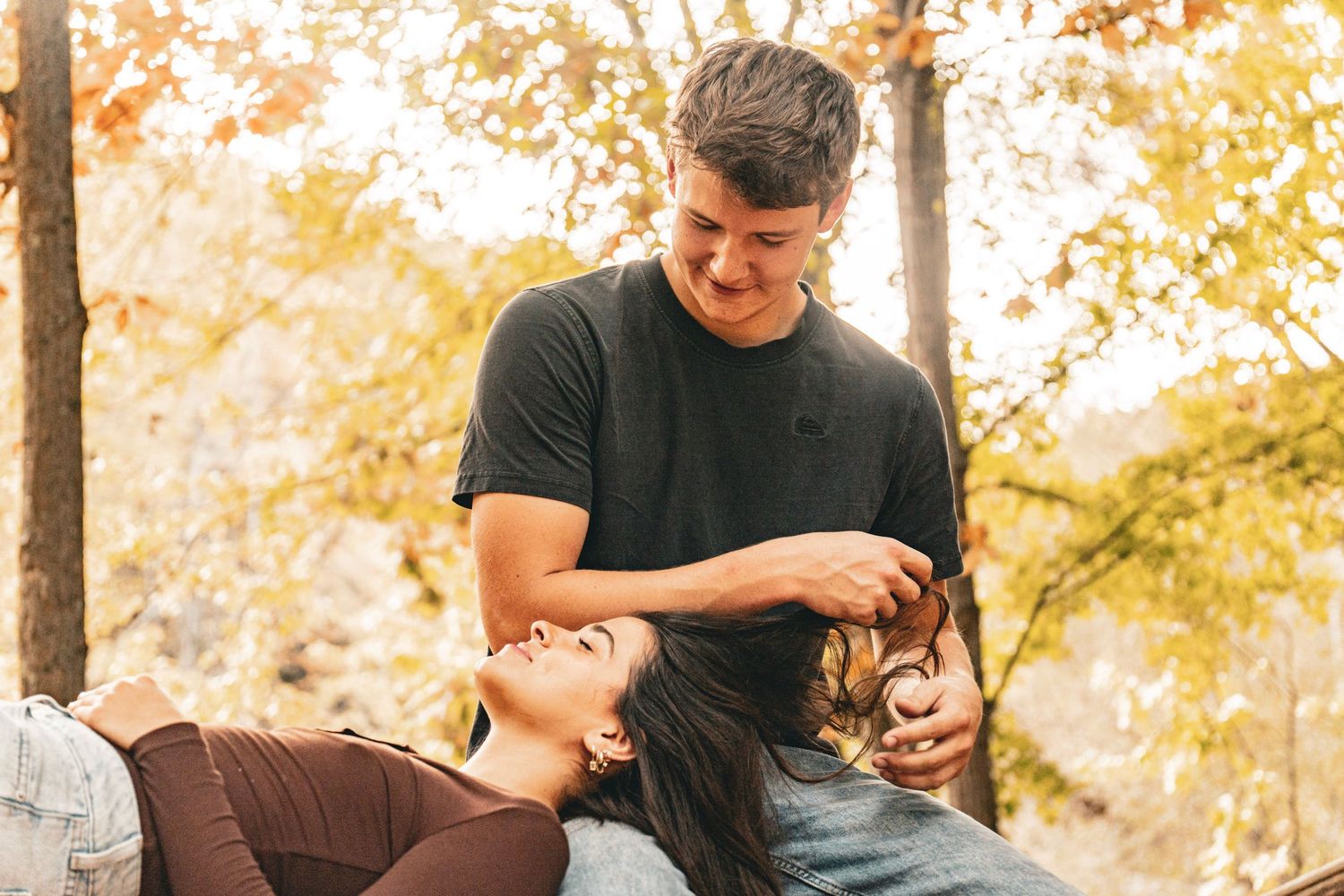 A smiling man in a dark t-shirt gently touches the hair of a woman lying in his lap amid golden autumn trees.