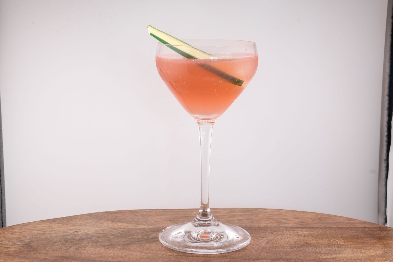 Elegant cocktail with orange-pink hue served in a stemmed coupe glass on wooden table.