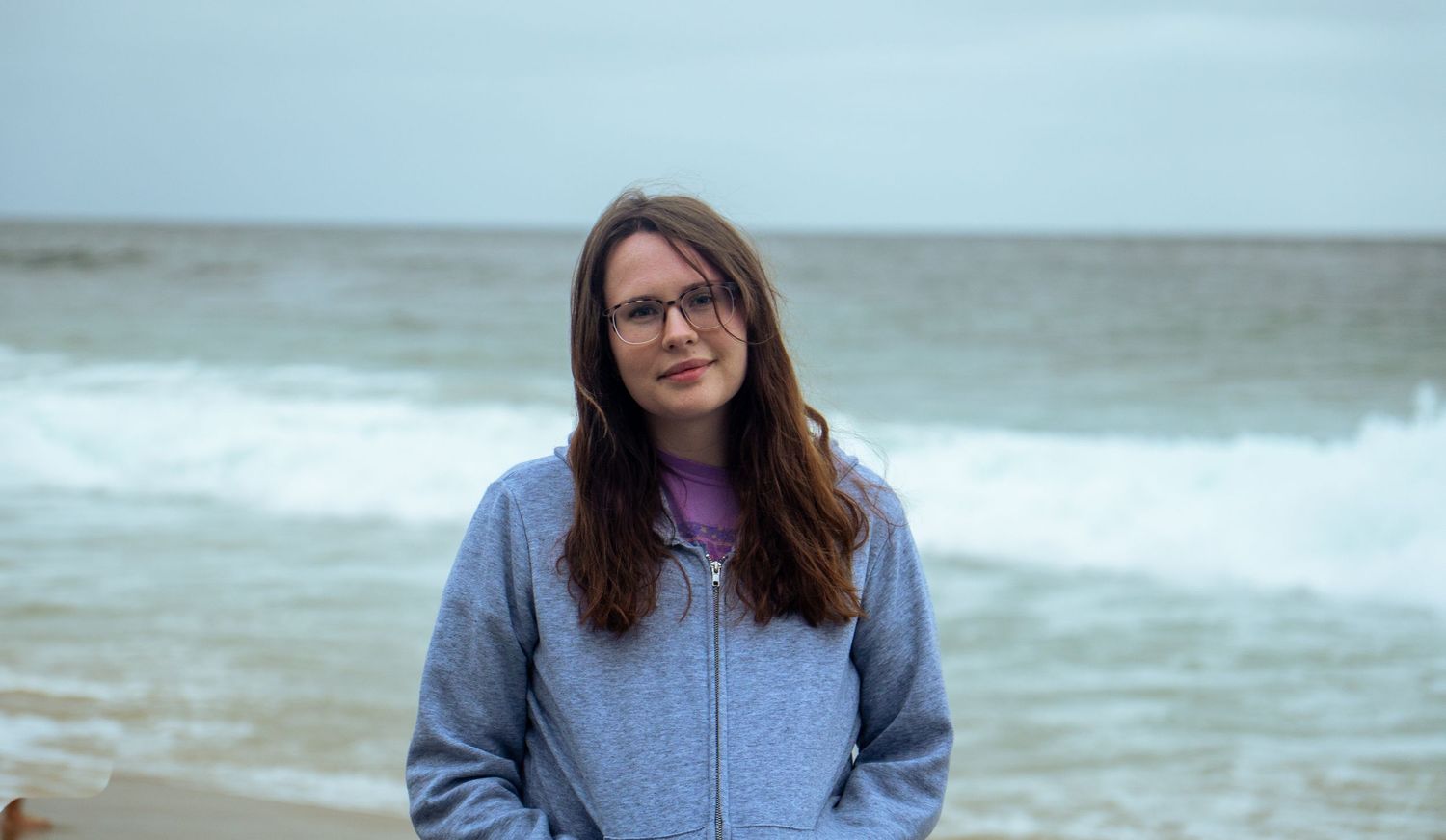 Casual beachgoer in glasses and gray sweatshirt poses by the ocean on an overcast day.
