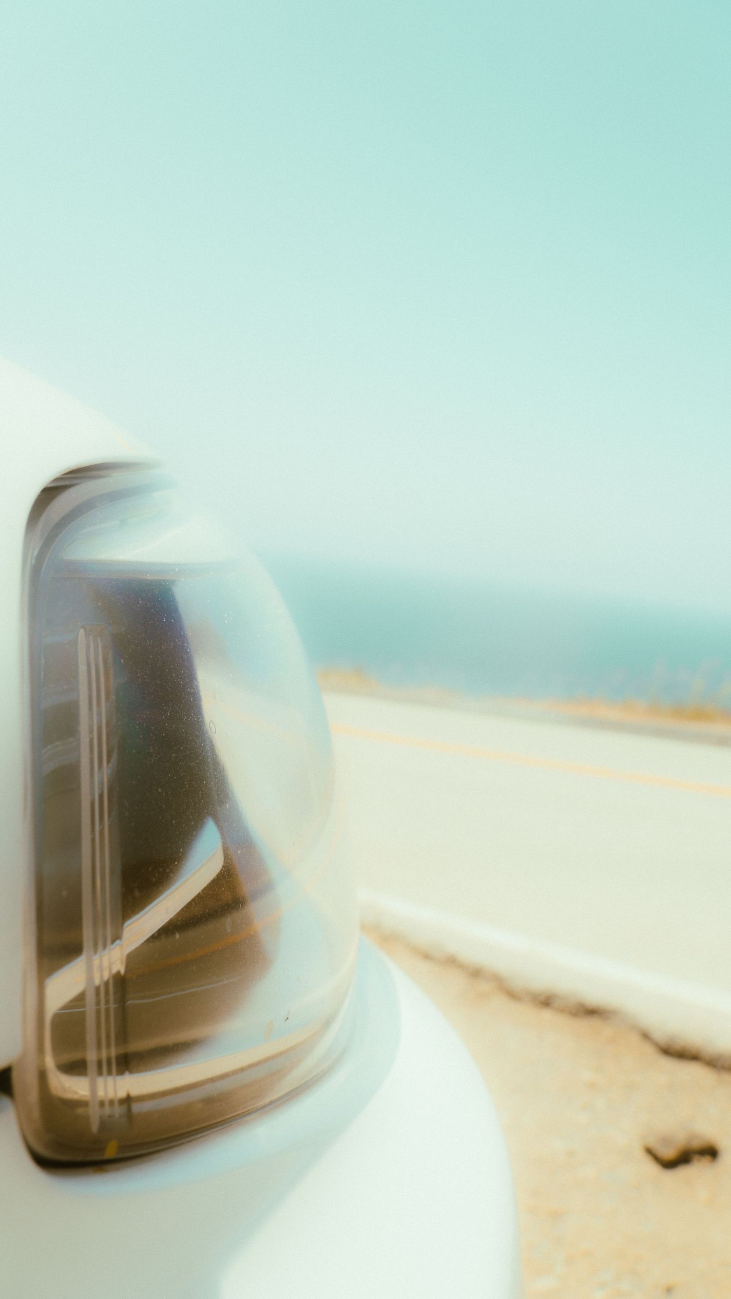Side mirror reflection of a coastal road with blurred turquoise ocean in background.