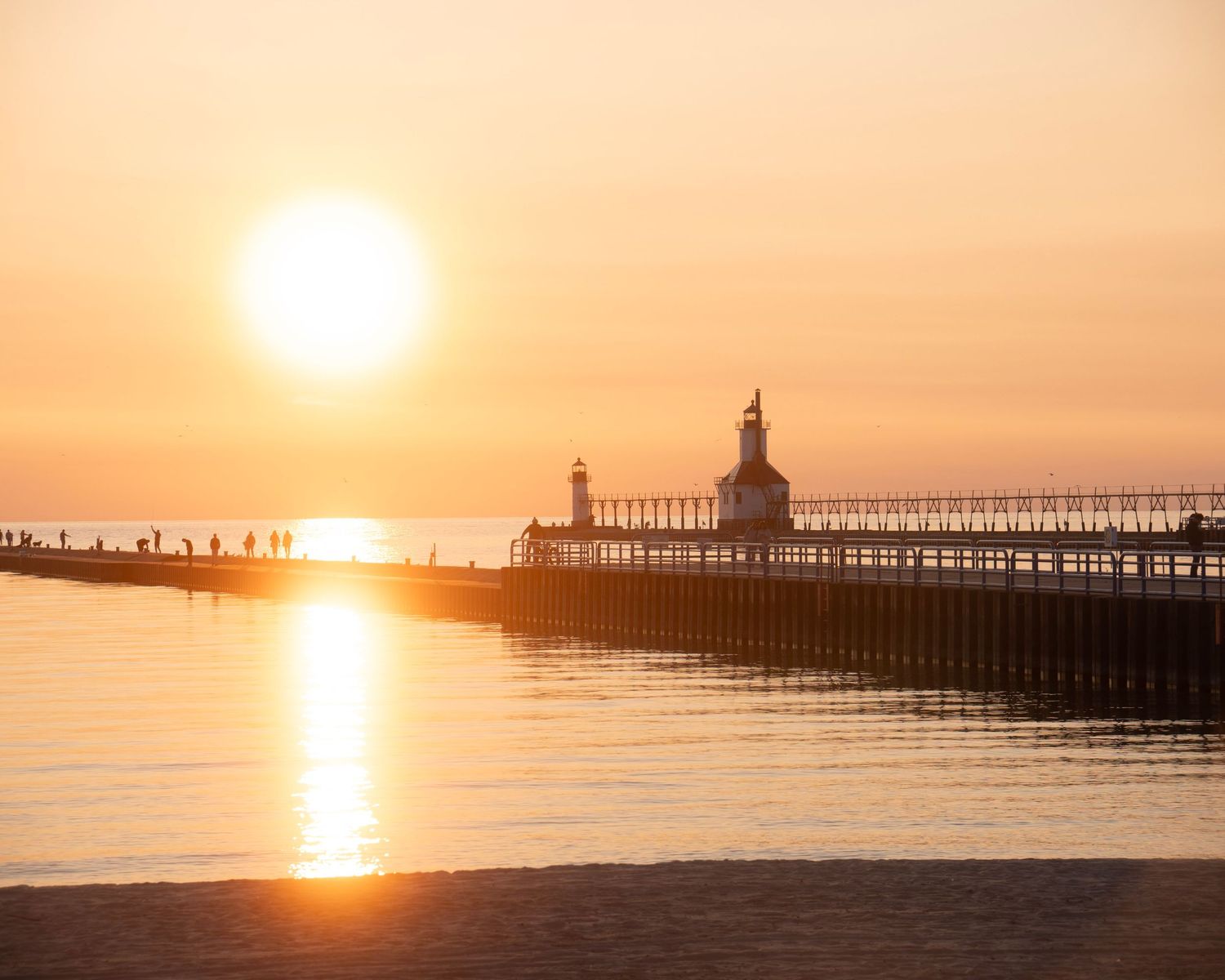 Lighthouse pier silhouetted against vibrant orange sunset over tranquil Lake Michigan waters.