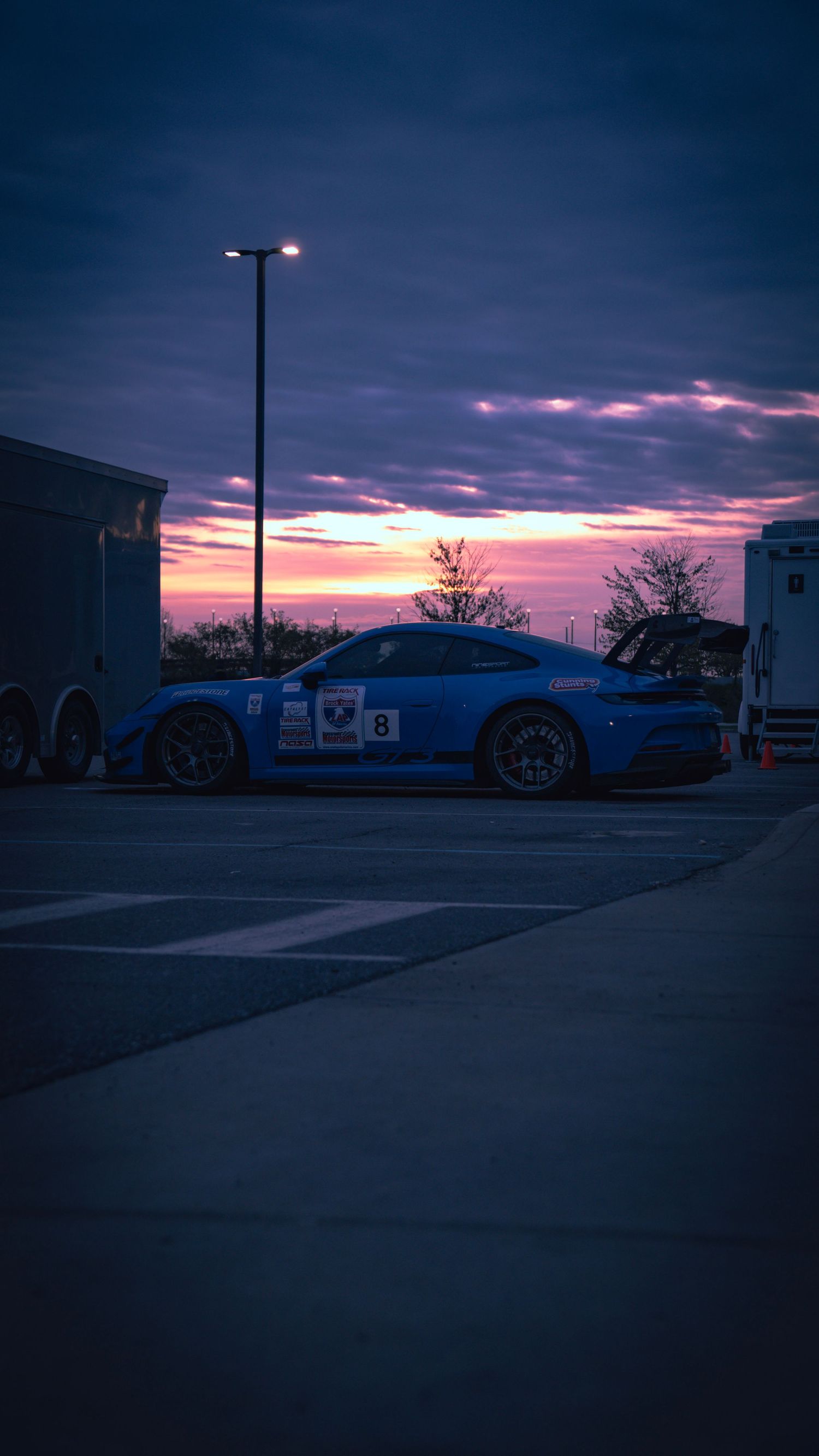 Blue sports car silhouetted against vibrant purple and pink sunset in parking lot.