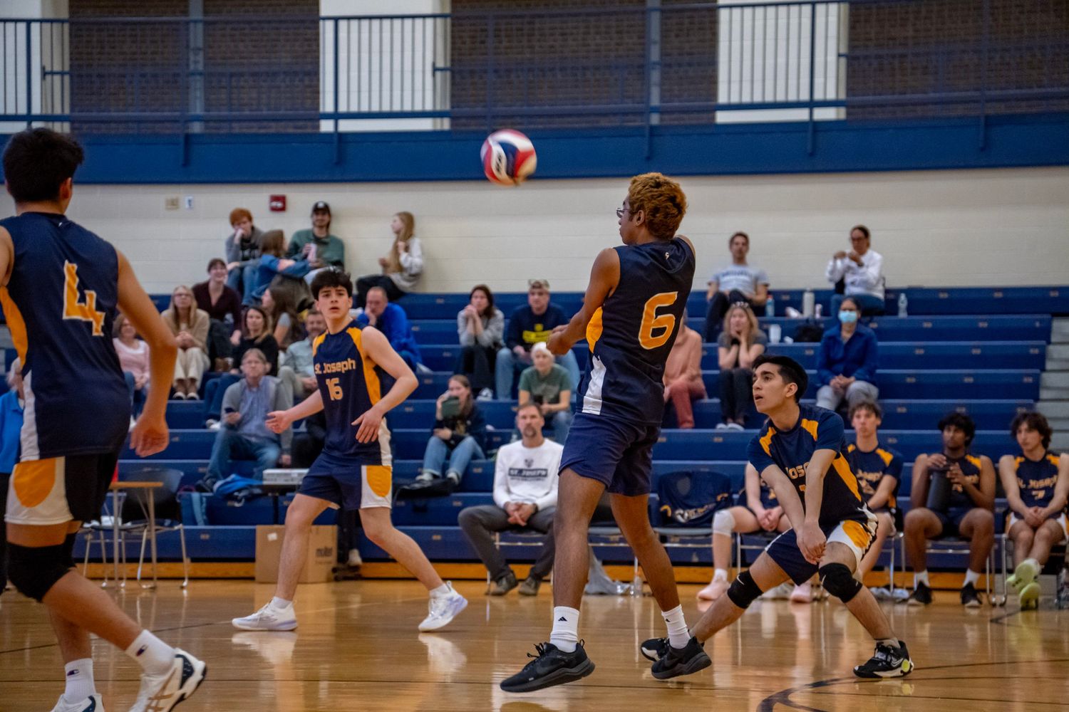 Indoor volleyball match in progress with players positioned around the court and spectators in the bleachers.