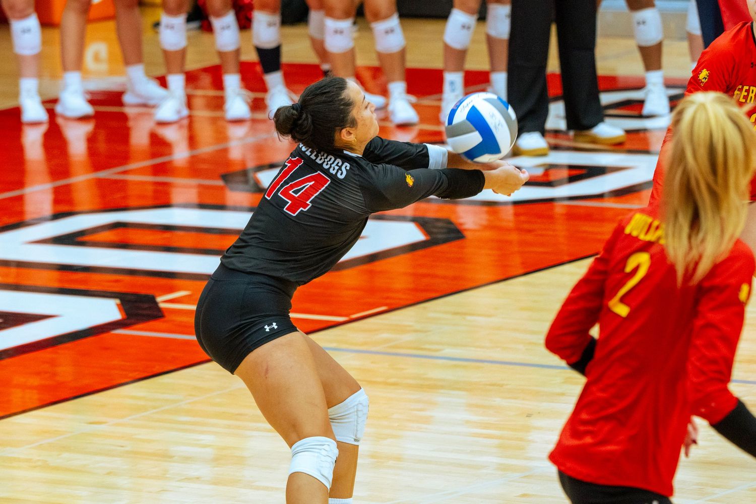 Volleyball player in black jersey dives to save the ball during a match on a bright red court.