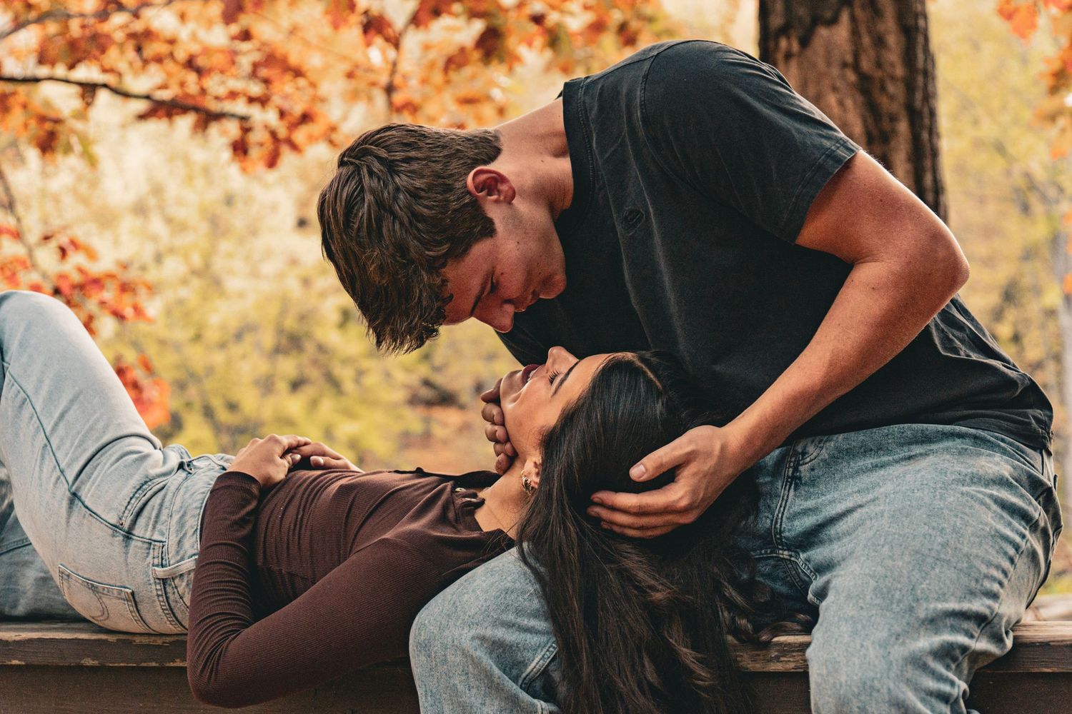 Couple sharing a tender moment on a wooden bench, a man leaning down to kiss a woman lying in his lap amid colorful autumn foliage.