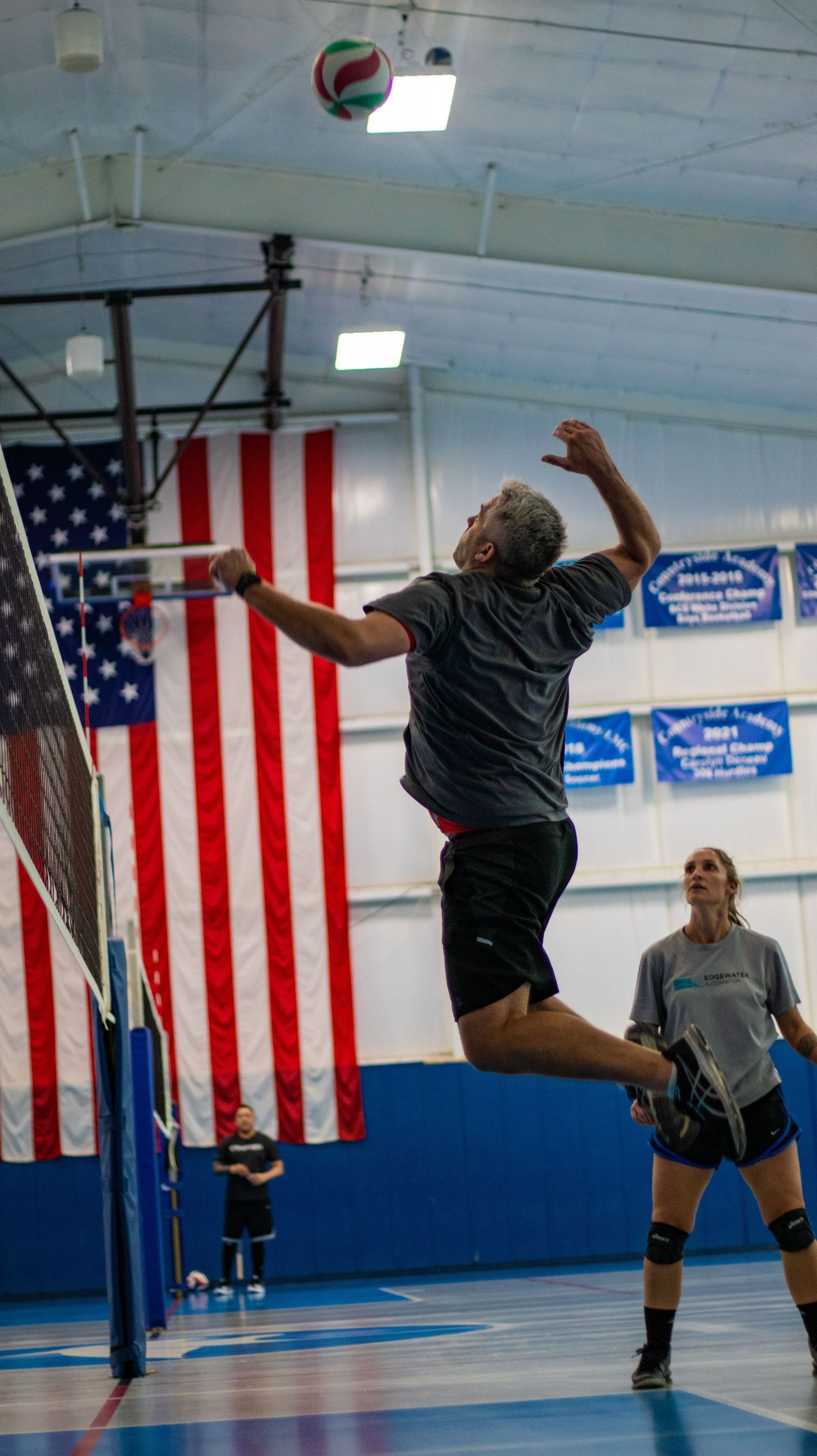 Volleyball player leaps high to spike ball during indoor match with American flag backdrop.