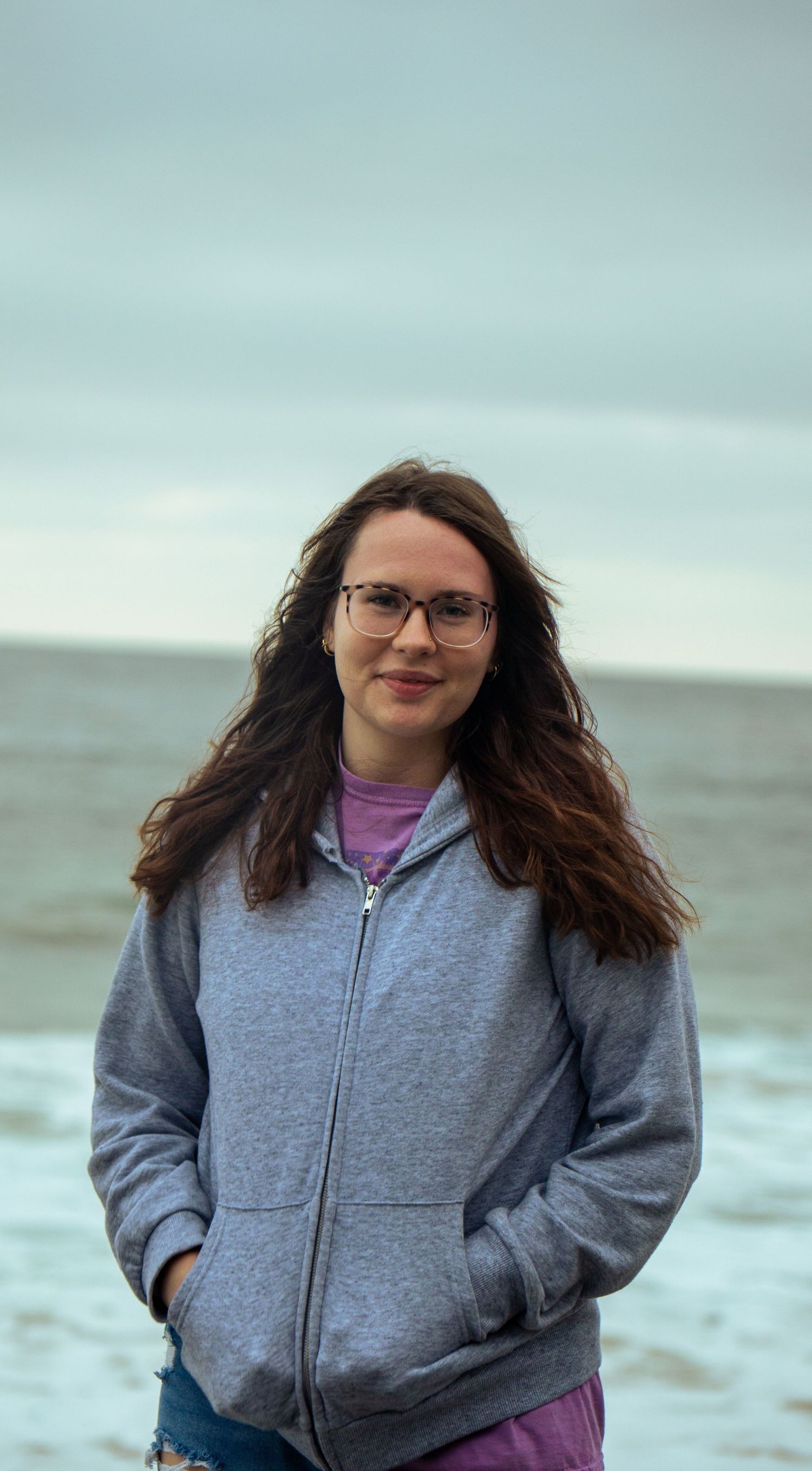 A relaxed beach portrait featuring someone in glasses and a gray zip-up hoodie against an ocean backdrop.