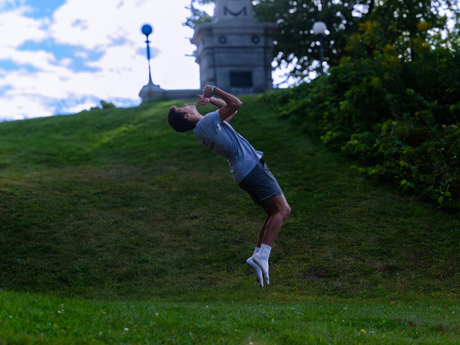 Athletic figure doing a backbend flip on a grassy hillside against blue sky.