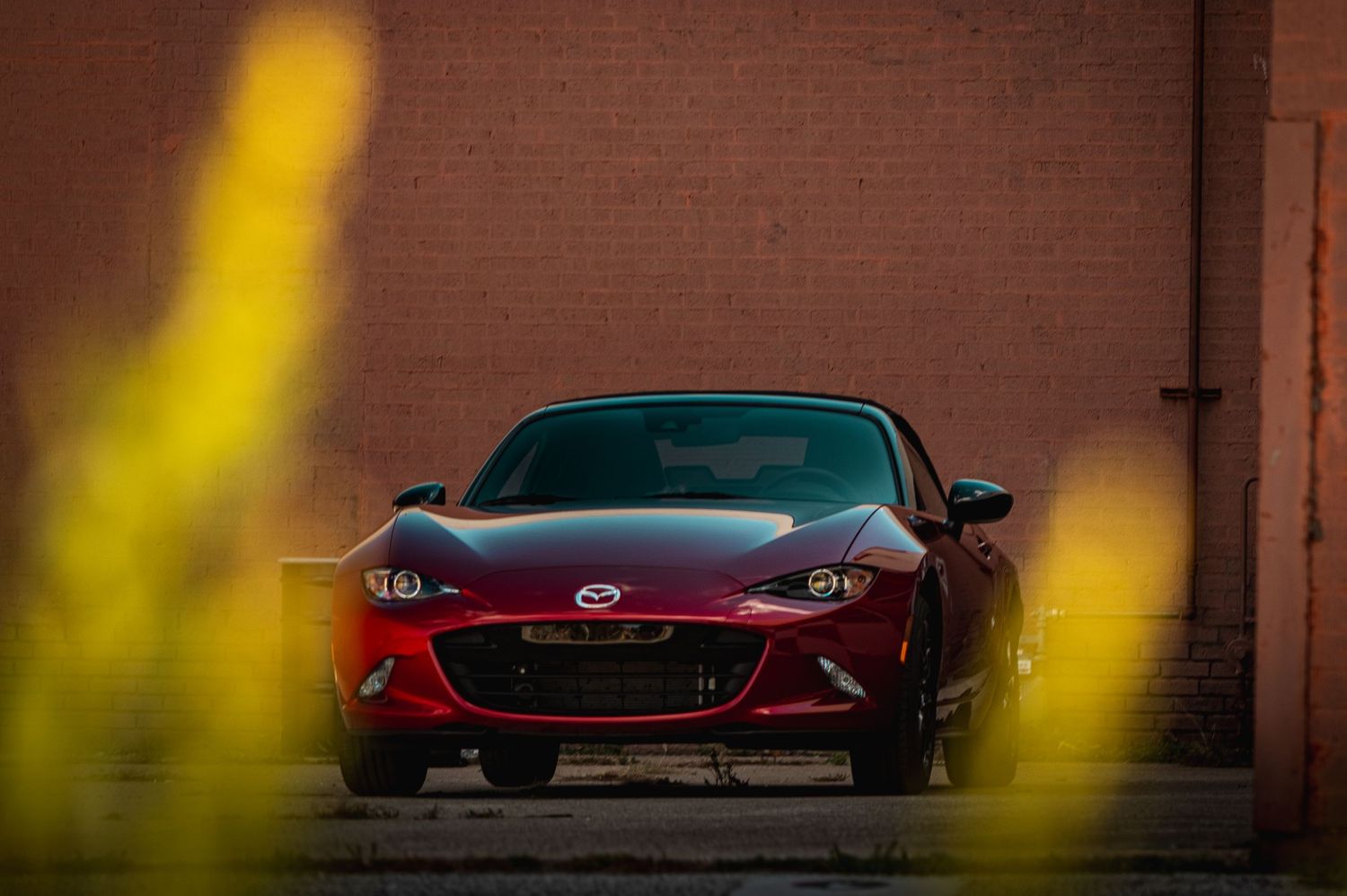 Front view of red Mazda MX-5 with yellow blur in foreground against brick wall.