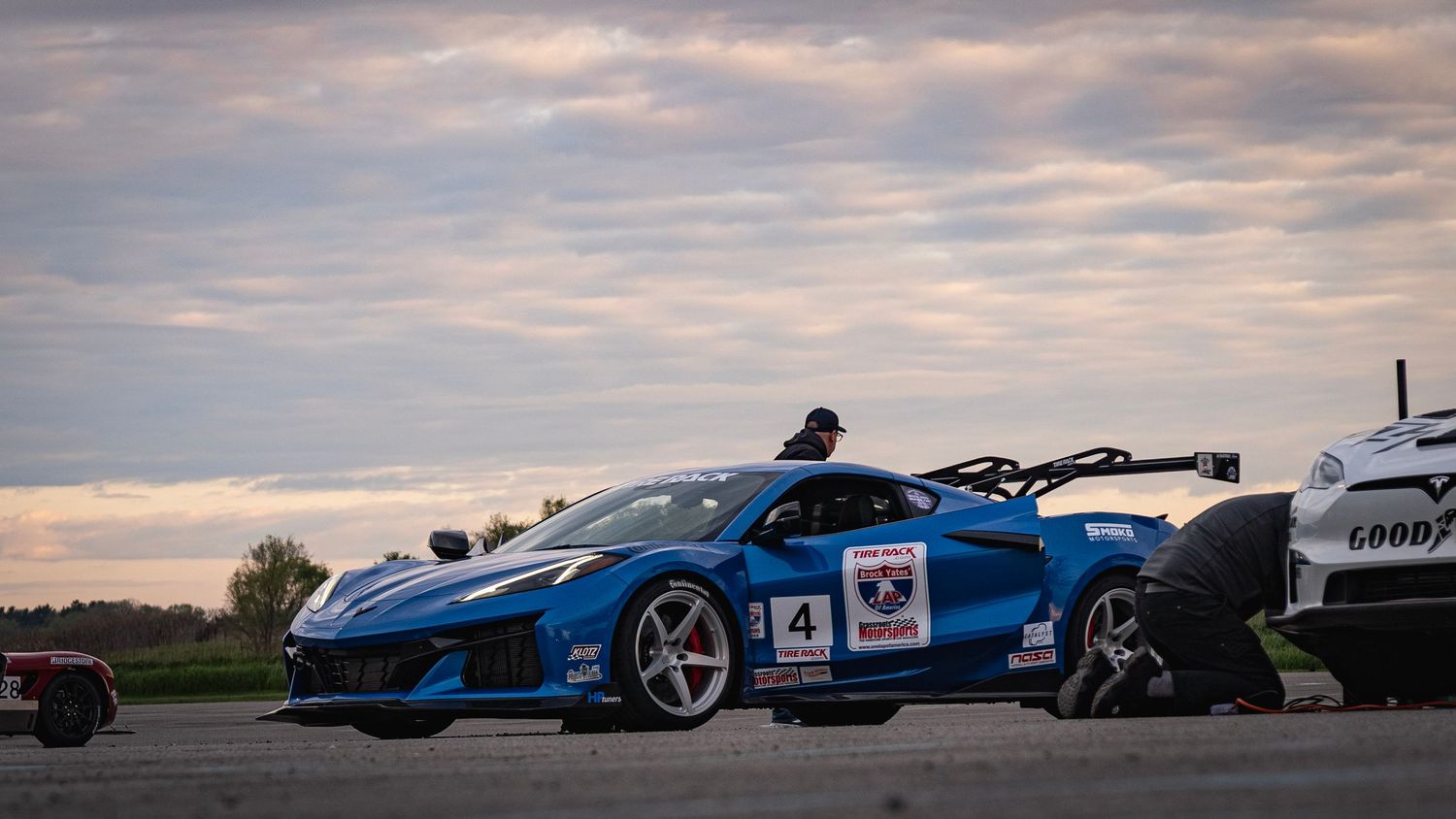 Blue race-modified sports car parked outdoors under cloudy sunset sky.