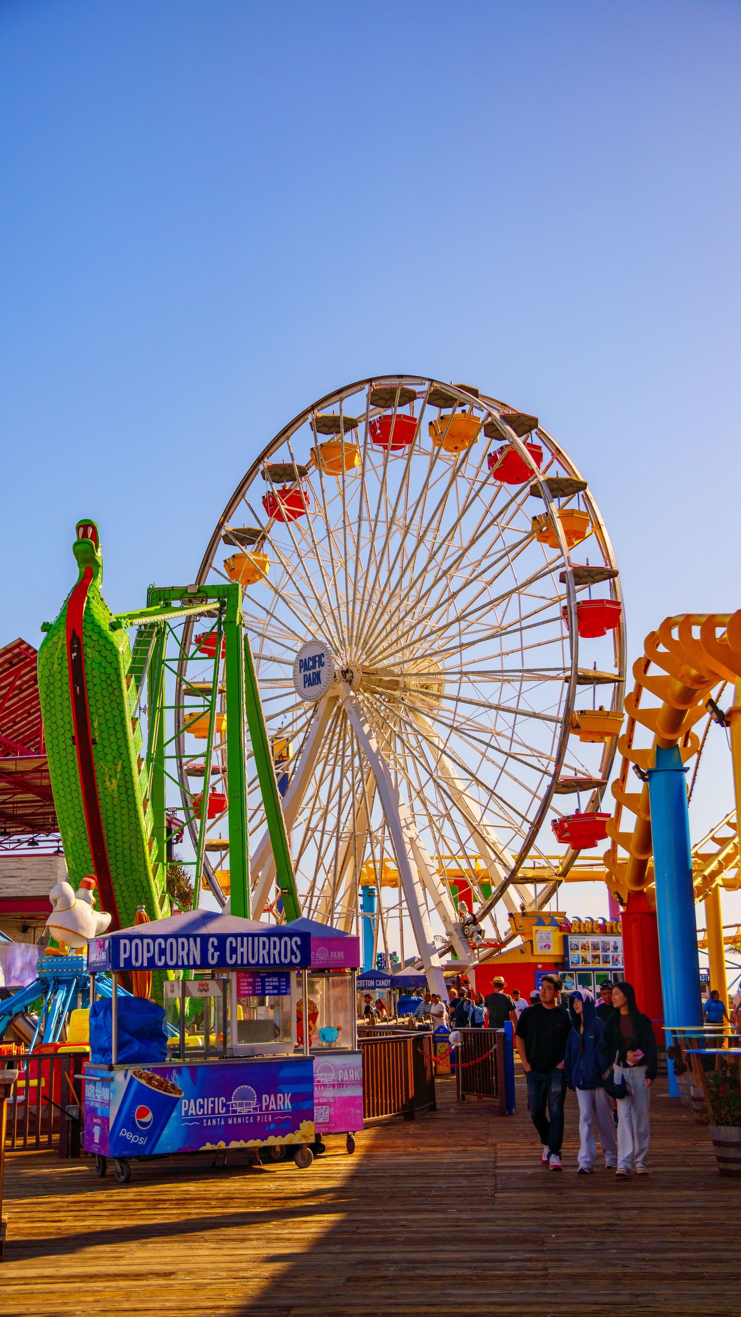 Colorful amusement park rides and Ferris wheel against clear blue sky on wooden pier boardwalk.