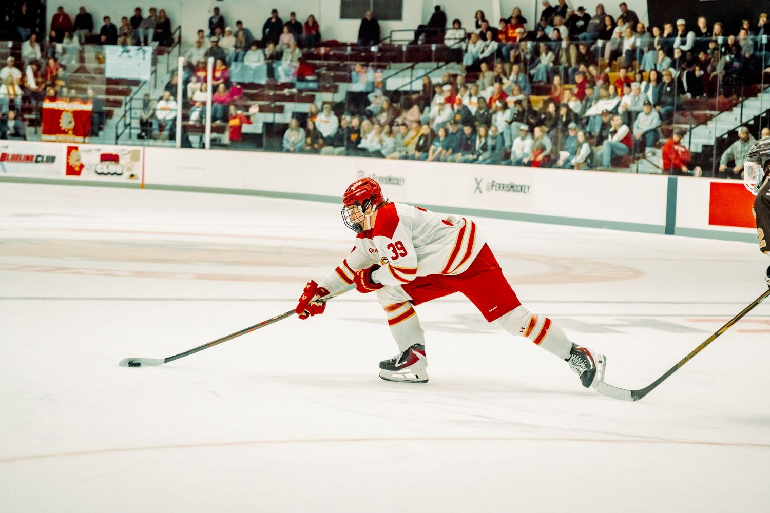 Hockey player in a red and white uniform and helmet glides on the ice with his stick extended during a game as fans watch from the stands.