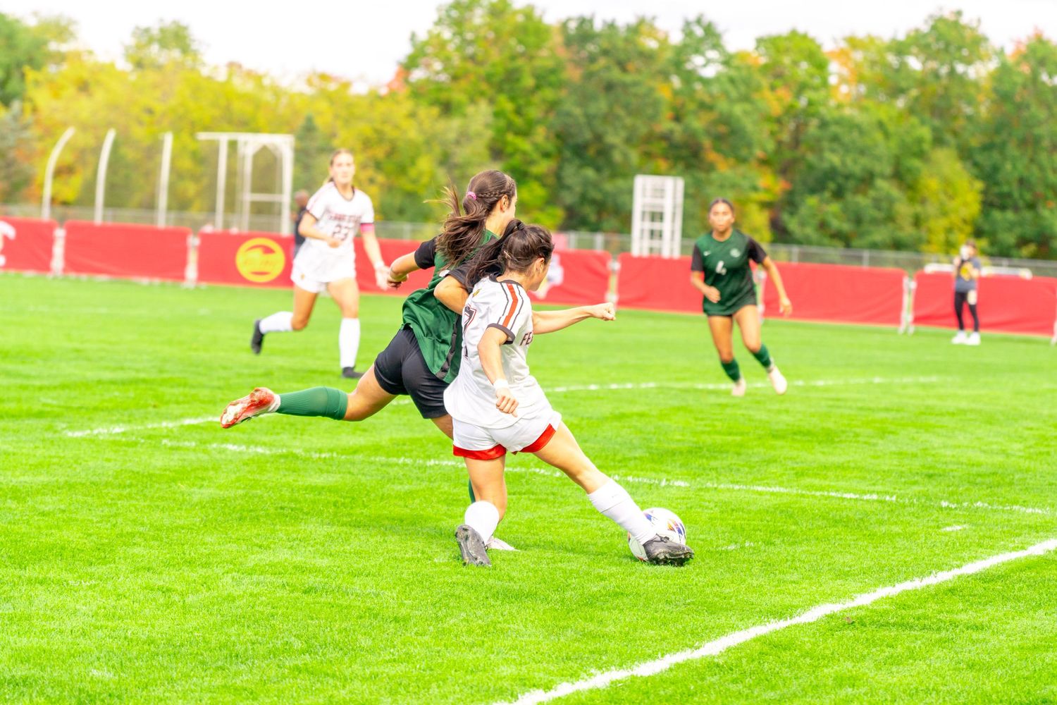 Female soccer players in white and green uniforms compete for the ball during a college match on a grassy field with trees behind.