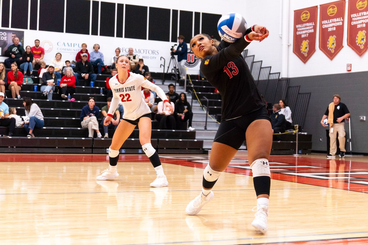 Volleyball player in a black jersey lunges to dig the ball during a college match while a teammate in white watches and fans look on.