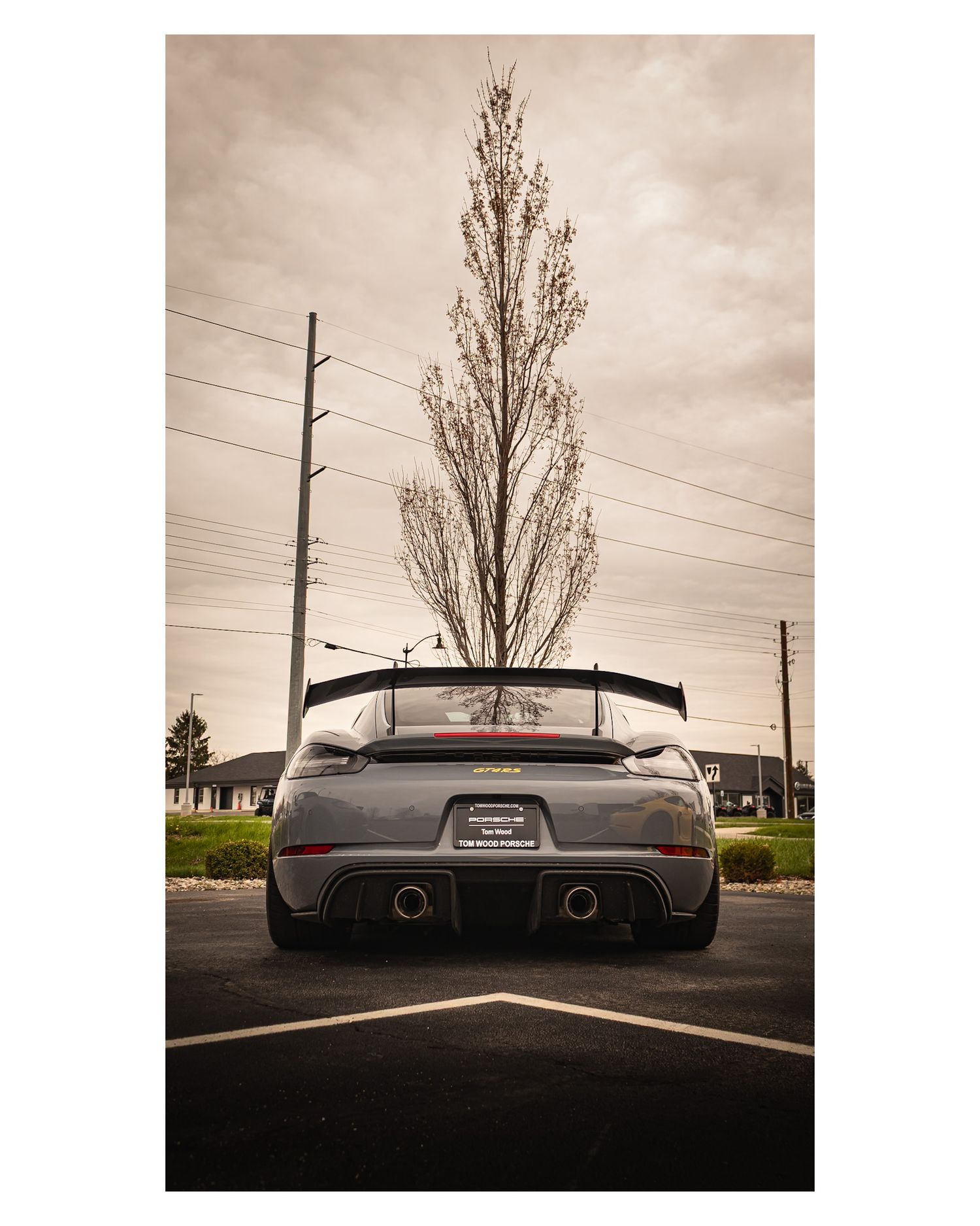 Monochrome rear view of sports car with large wing silhouetted against cloudy sky and utility poles.