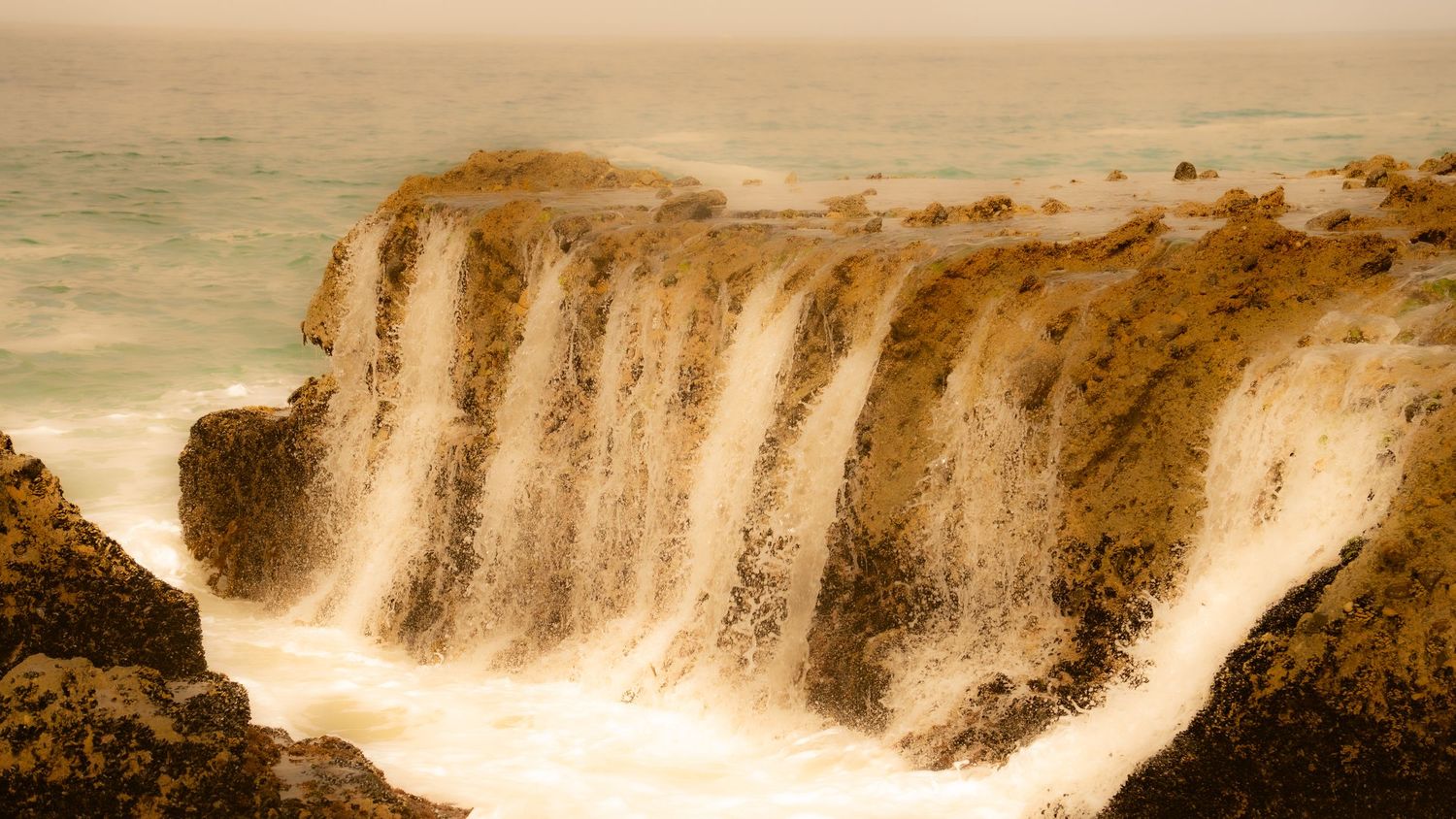 Dramatic sepia-toned waterfall cascades down steep coastal cliffs into the ocean.