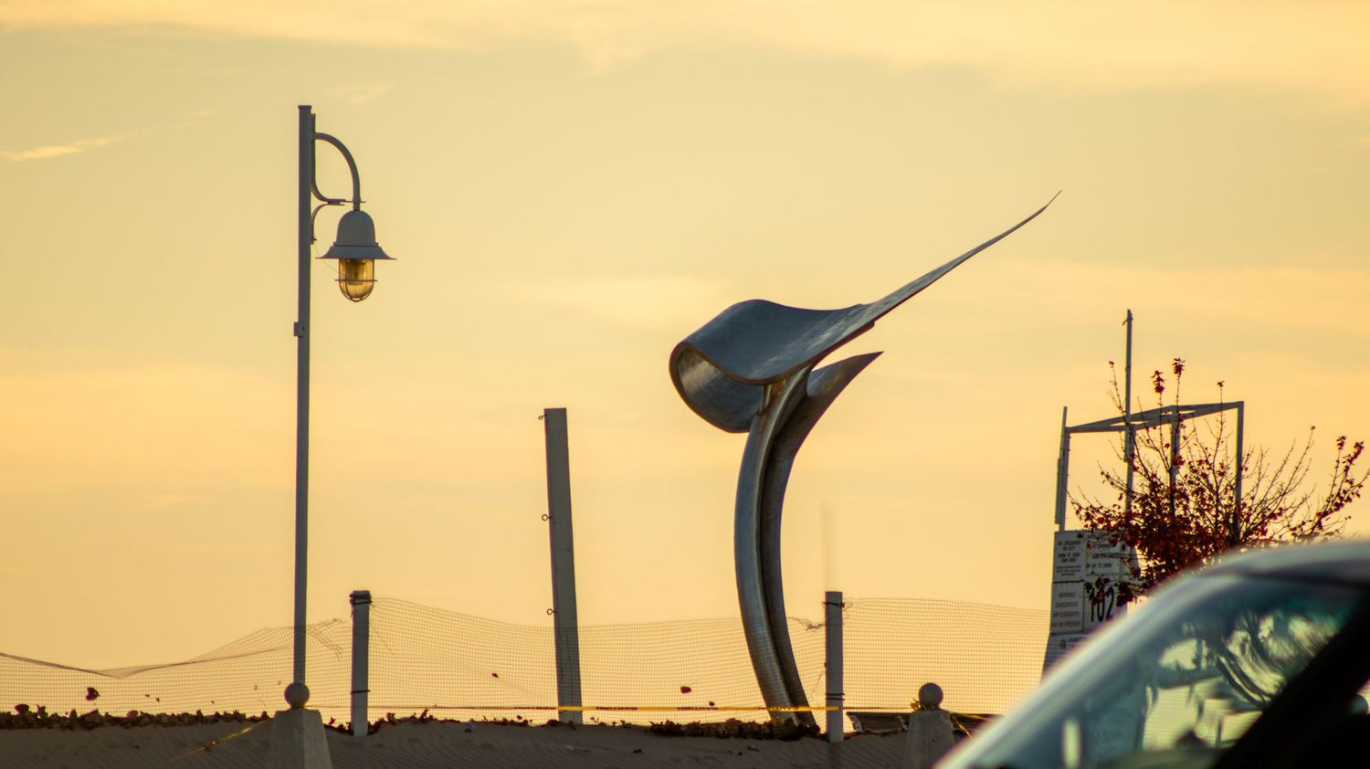 Modern sculptural marlin statue silhouetted against a golden sunset sky with a street lamp in foreground.