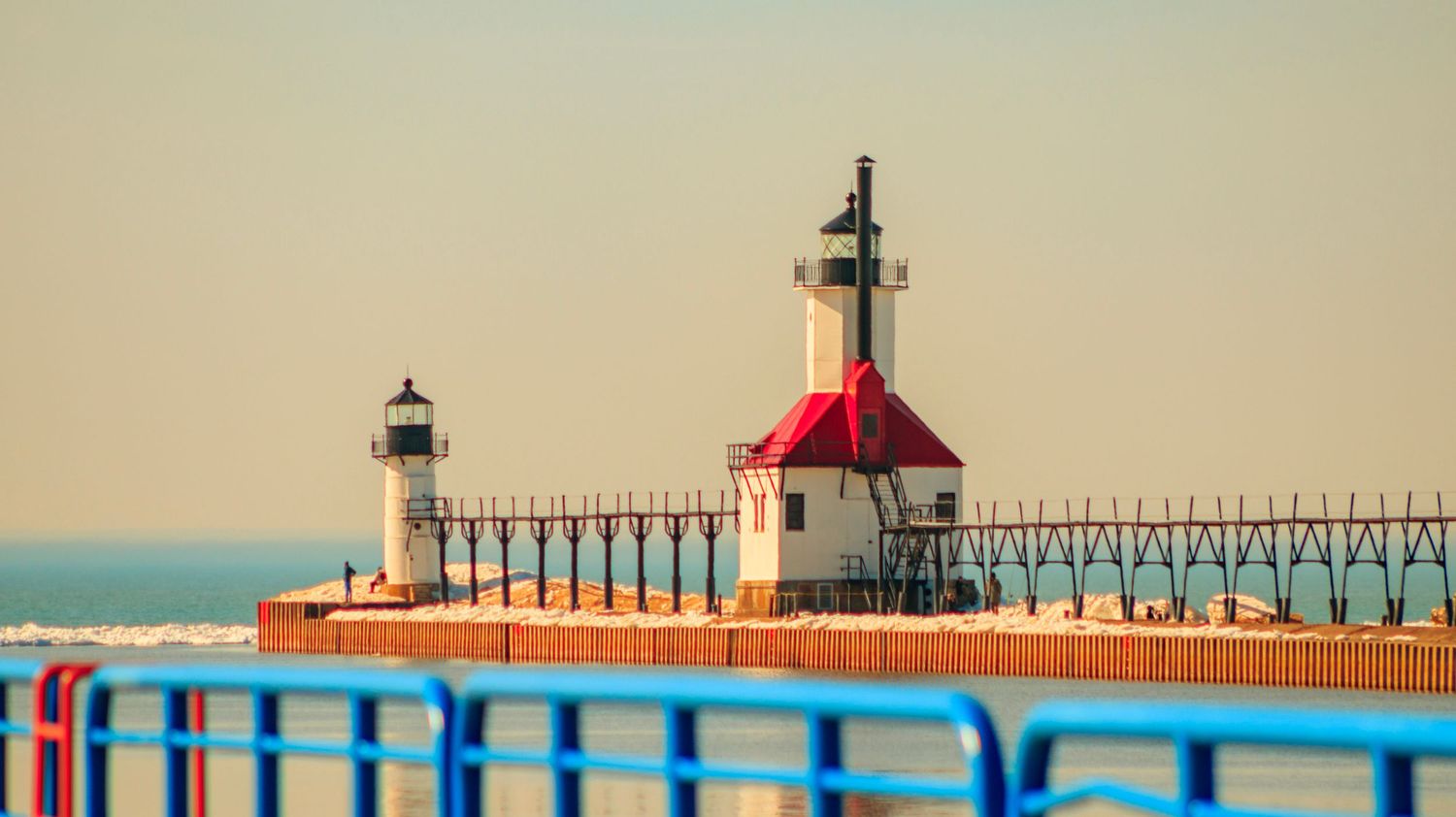 Red-capped lighthouse with long pier captured in warm tones against calm waters.