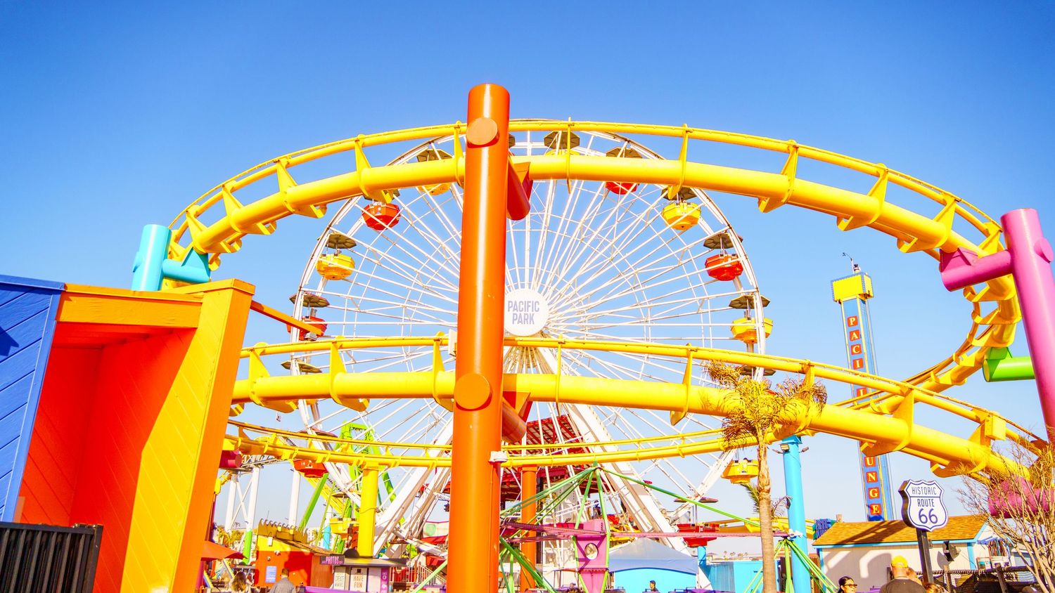 Panoramic view of amusement park rides and attractions with a Ferris wheel against a bright blue sky.