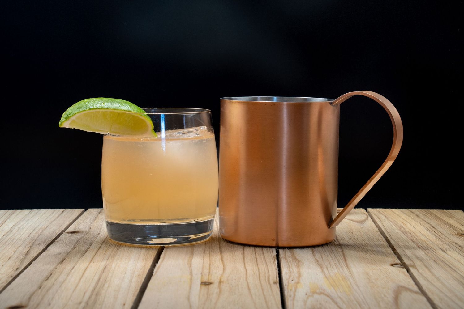 Citrus cocktail with lime garnish beside copper mug on rustic wooden table.