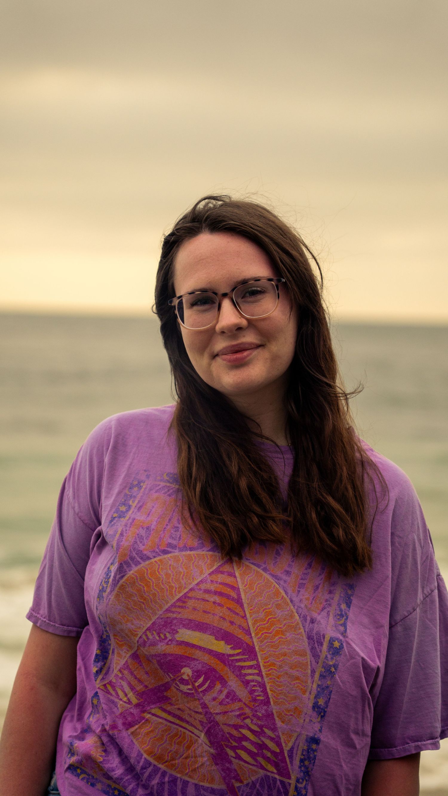 Person in purple t-shirt smiles during golden hour at beach with soft lighting.
