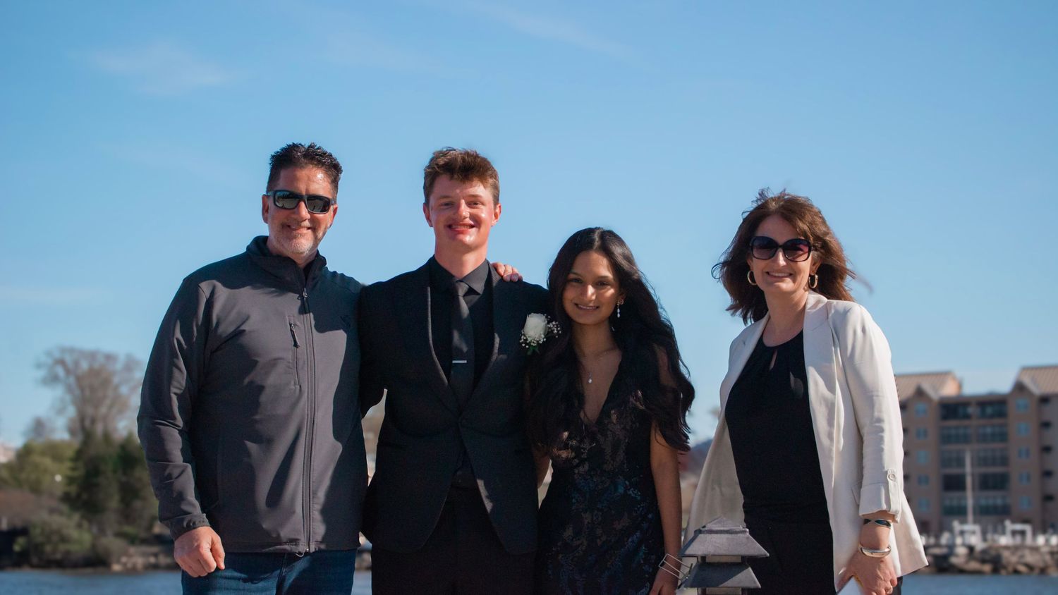 A group of four people standing together outdoors on a sunny day with blue skies and water in the background.