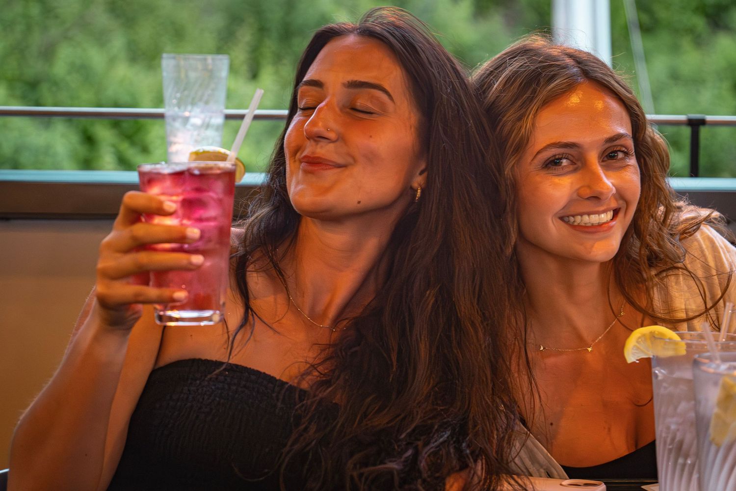 Two women with long brown hair enjoying colorful cocktails together while seated at a dining establishment.