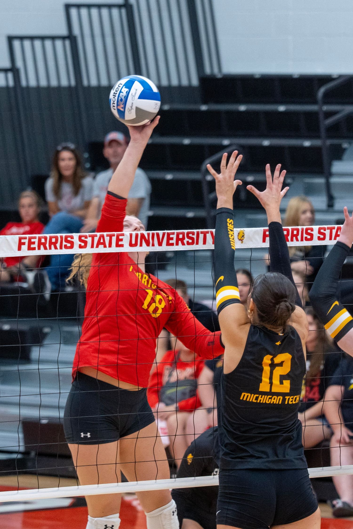 Volleyball players from Ferris State and Michigan Tech compete at the net during an intense match.