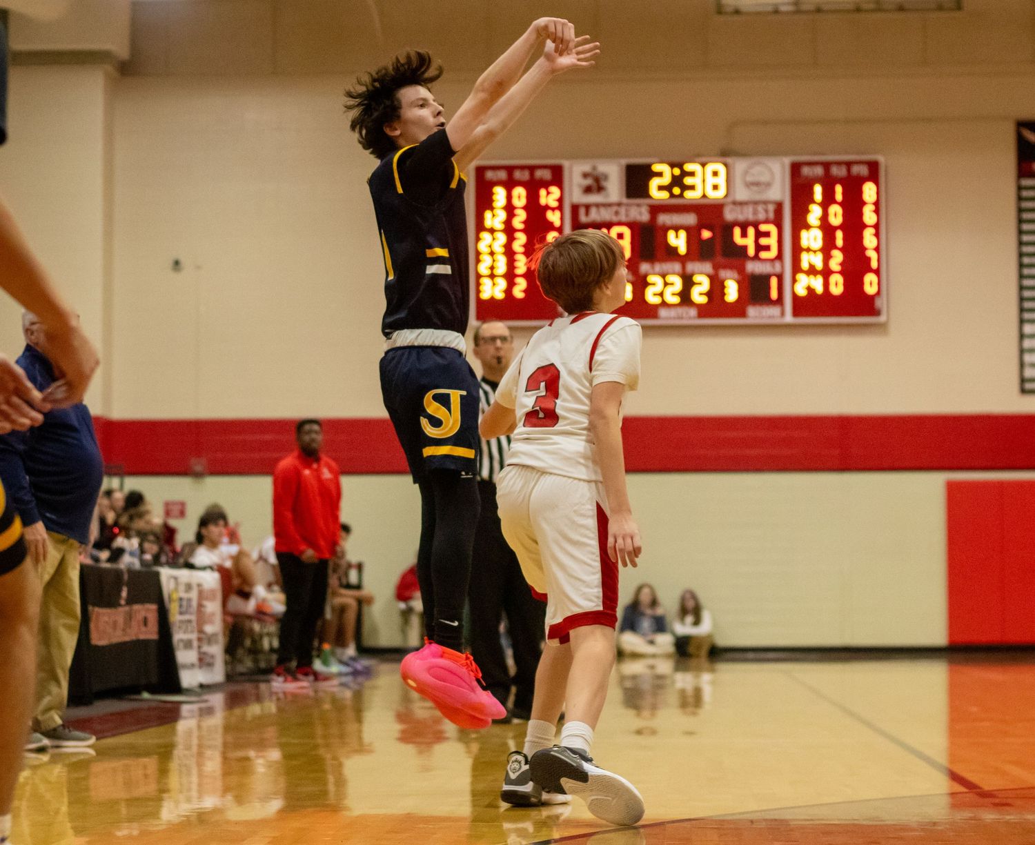 Player in dark jersey takes a jump shot over defender in white uniform with scoreboard visible in background.