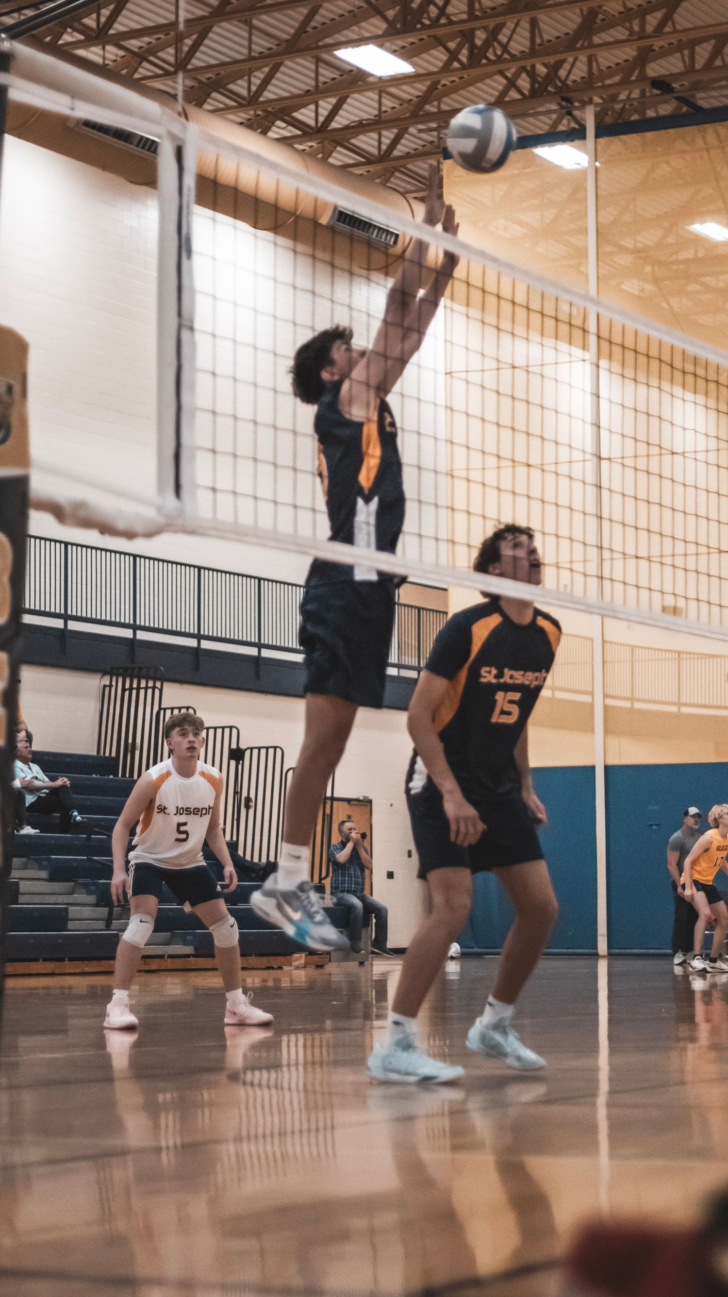 A volleyball player reaches high to block the ball at the net during an indoor game.
