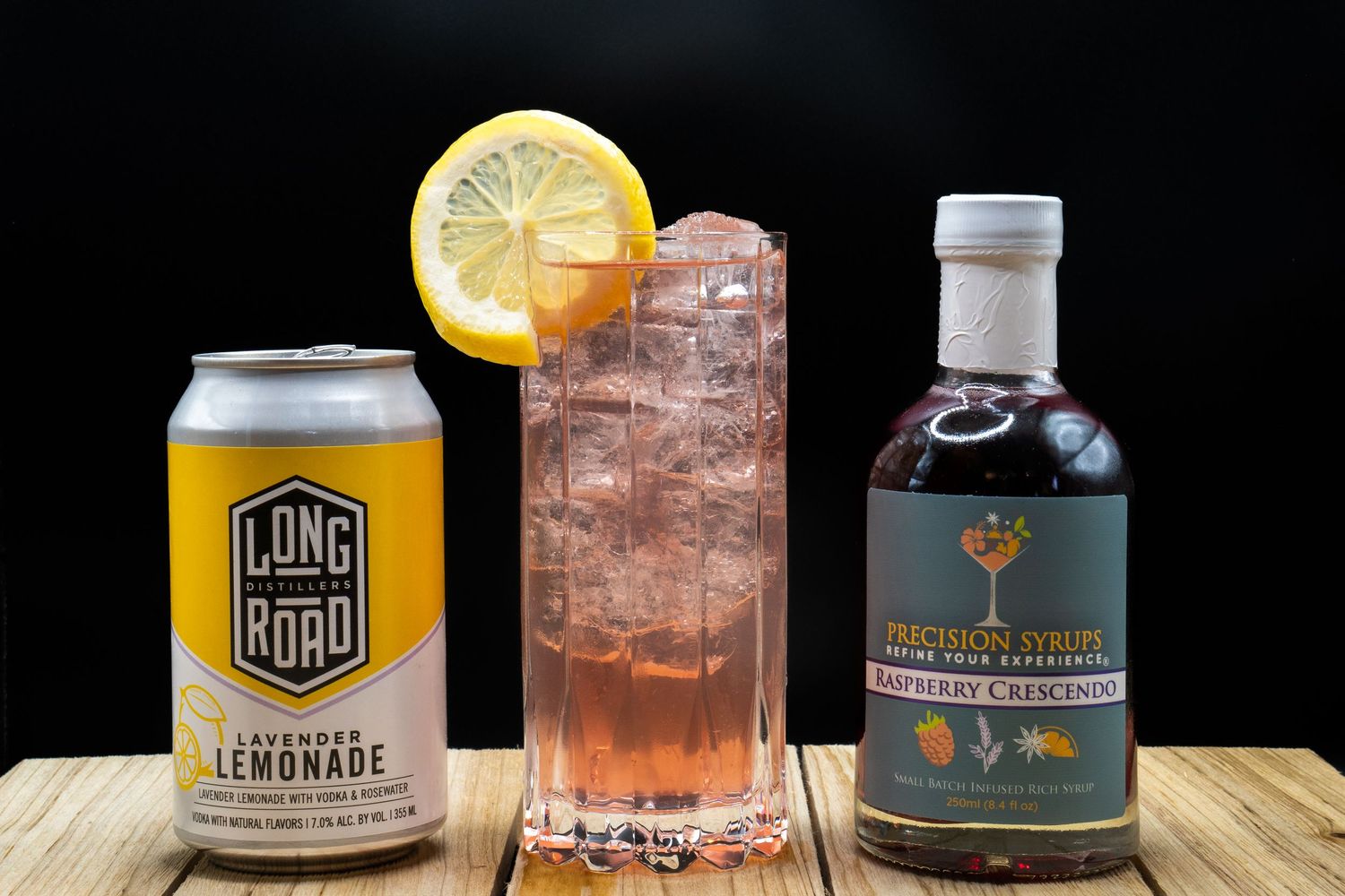 Pink highball cocktail with lemon garnish flanked by Long Road can and Precision Syrup bottle on wooden surface.