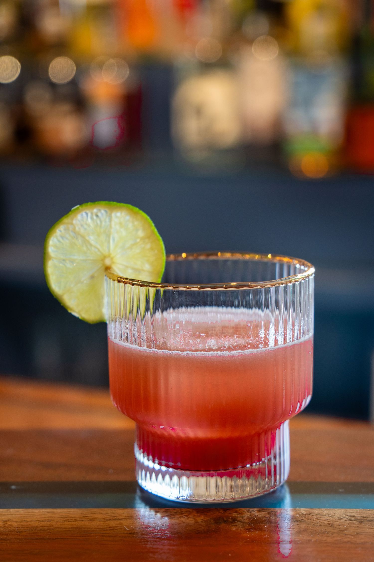 A red craft cocktail with lime garnish in a ribbed coupe glass on a wooden bar surface.