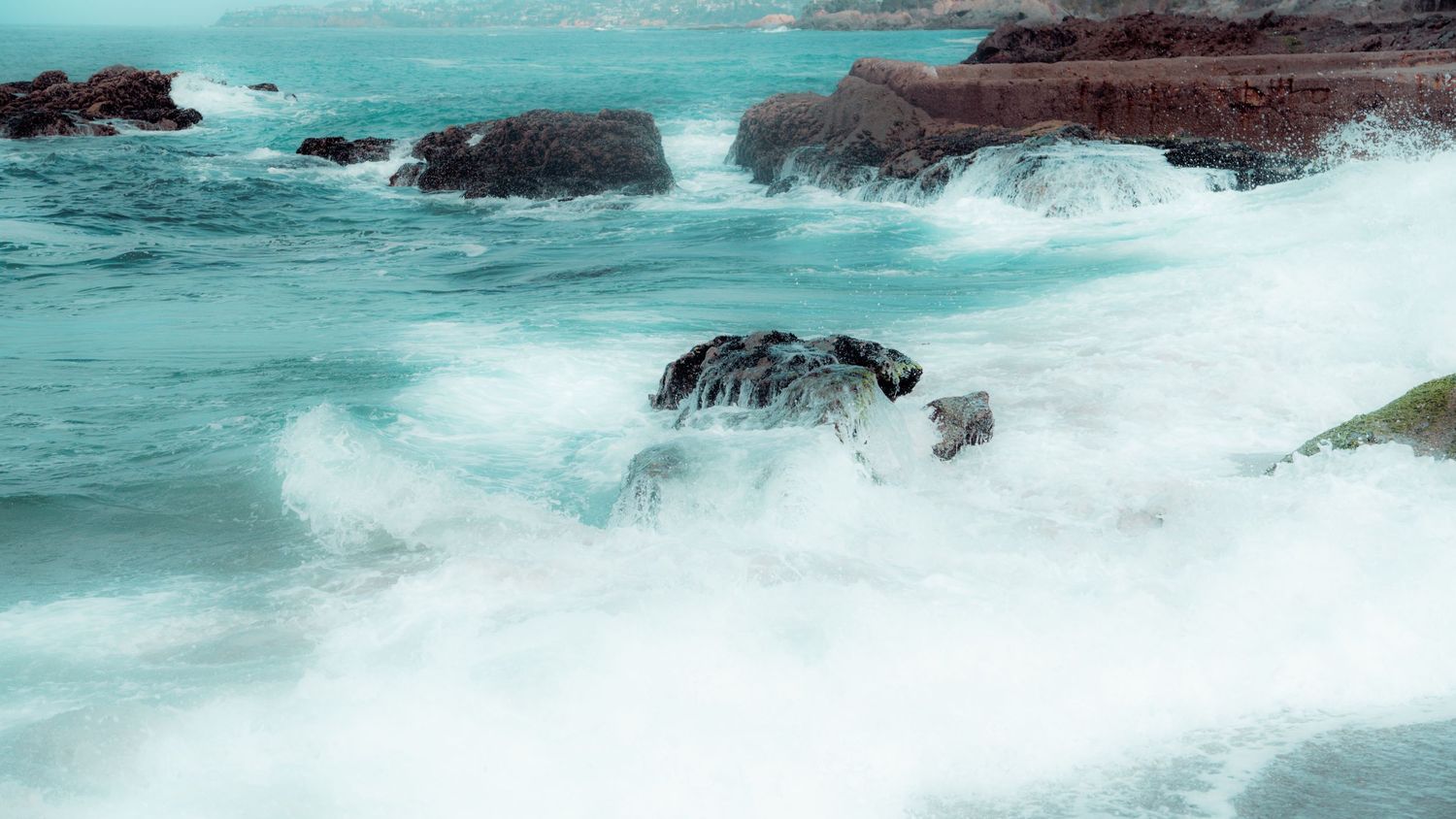Powerful waves crash against dark rocky outcrops in turquoise ocean waters creating dramatic white spray.