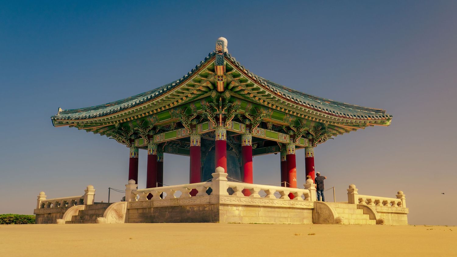 Traditional Korean pavilion with green curved roof and red pillars standing in desert-like landscape.