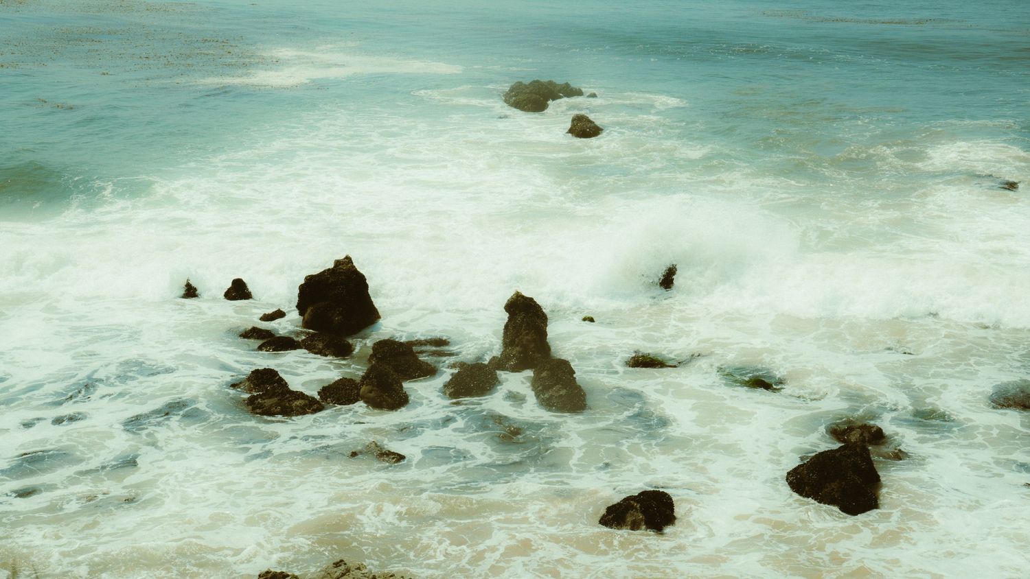 Dramatic seascape of waves crashing against scattered rock formations in turquoise waters.