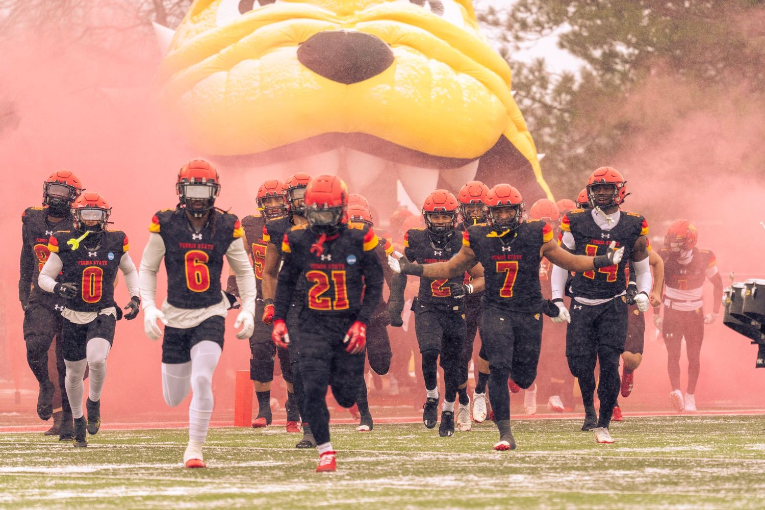 Football players run through orange smoke during a dramatic game entrance ceremony at a sports field.