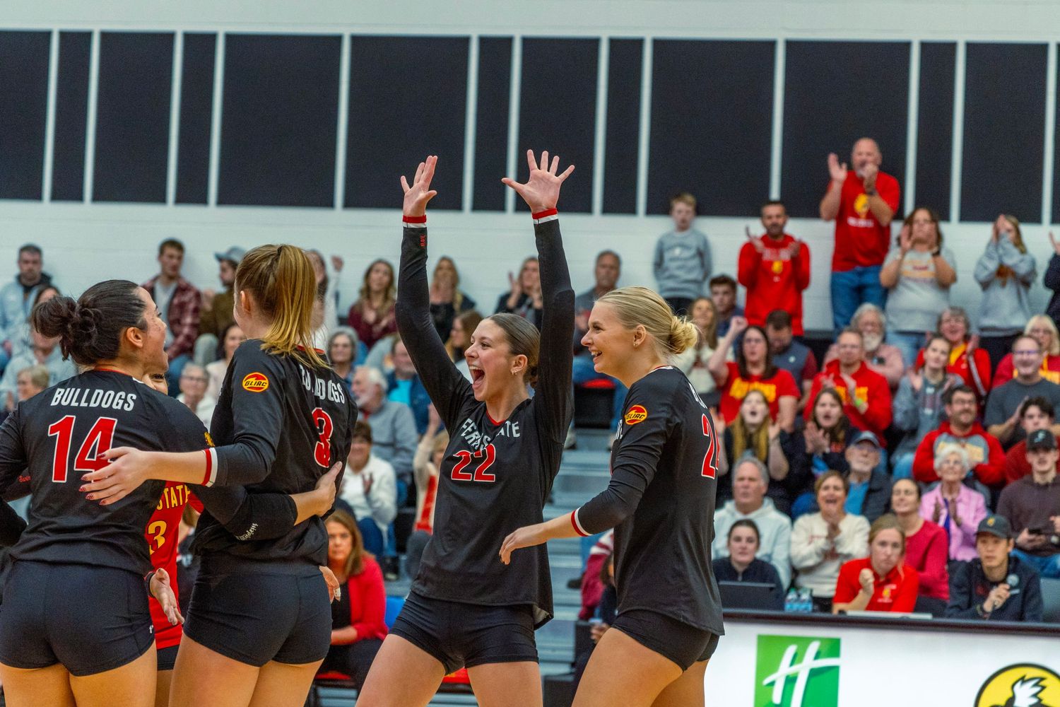 Volleyball players celebrate and jump with excitement during a match while spectators watch from the stands.