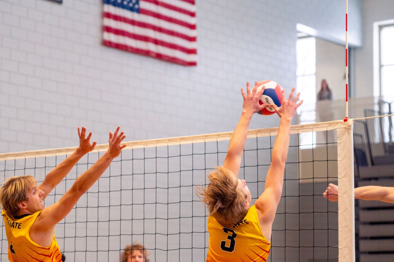 Volleyball players leaping at the net during an intense match with American flag visible in background.