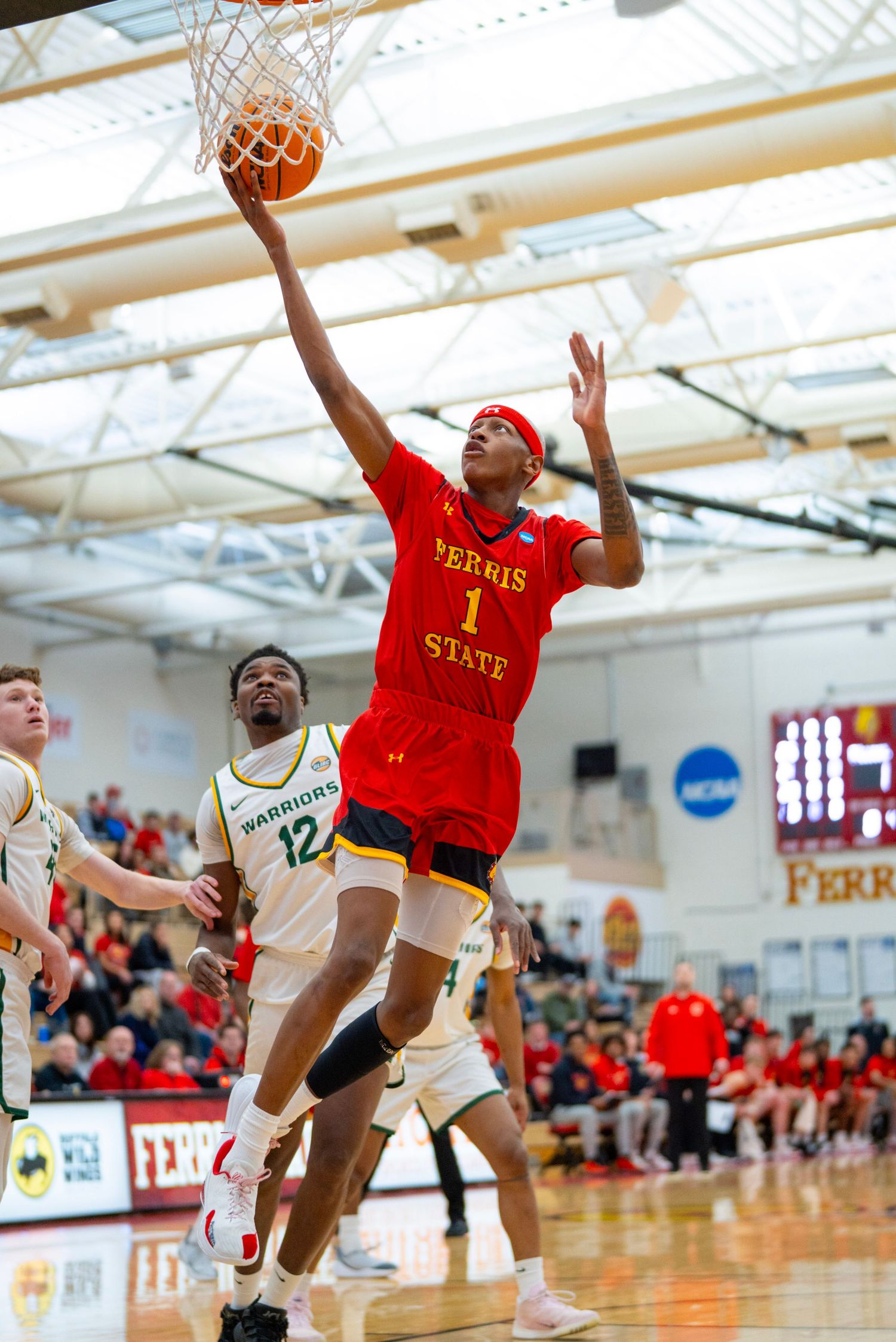 Basketball player in red jersey leaps high to make a layup during an intense indoor game at a college gymnasium.