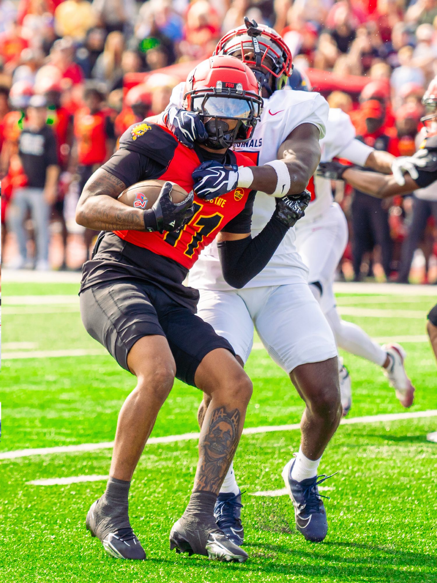 A football player in a red helmet and jersey carries the ball while a defender in white grabs him during a game in a packed stadium.