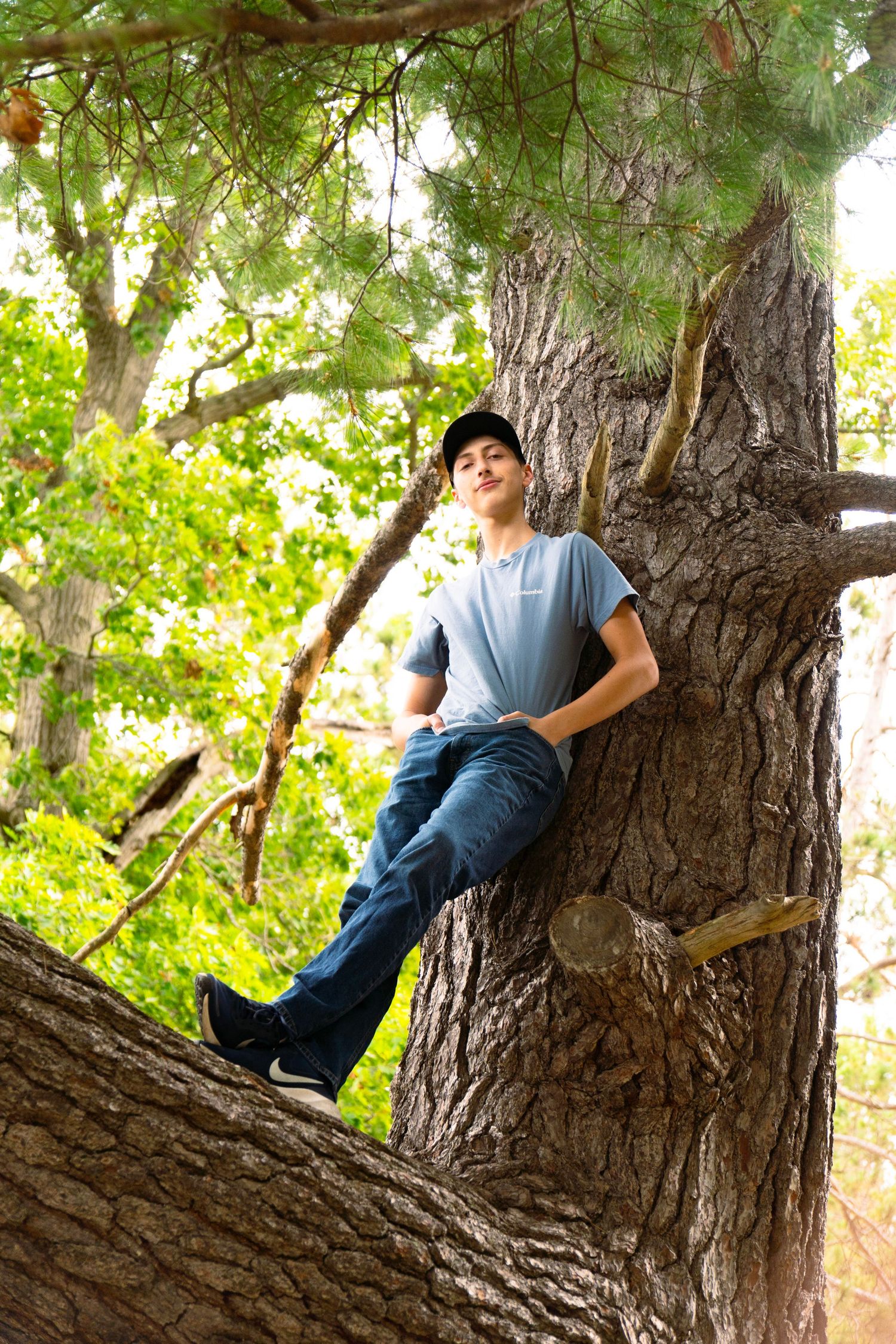 Young man in a cap, blue t-shirt and jeans sits relaxed on a thick tree branch, surrounded by green leaves and dappled sunlight.